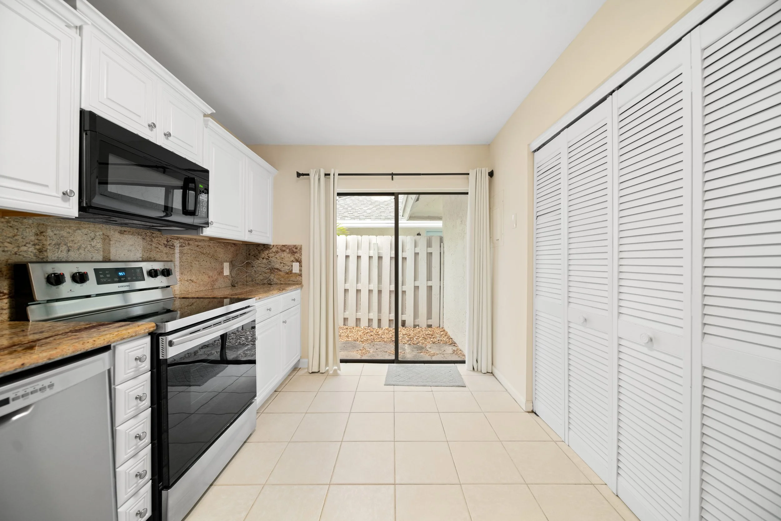 A kitchen with white cabinets, black and stainless stove, microwave, beige tiled floor, and a sliding glass door with white curtains leading to an outdoor area with a wooden fence.