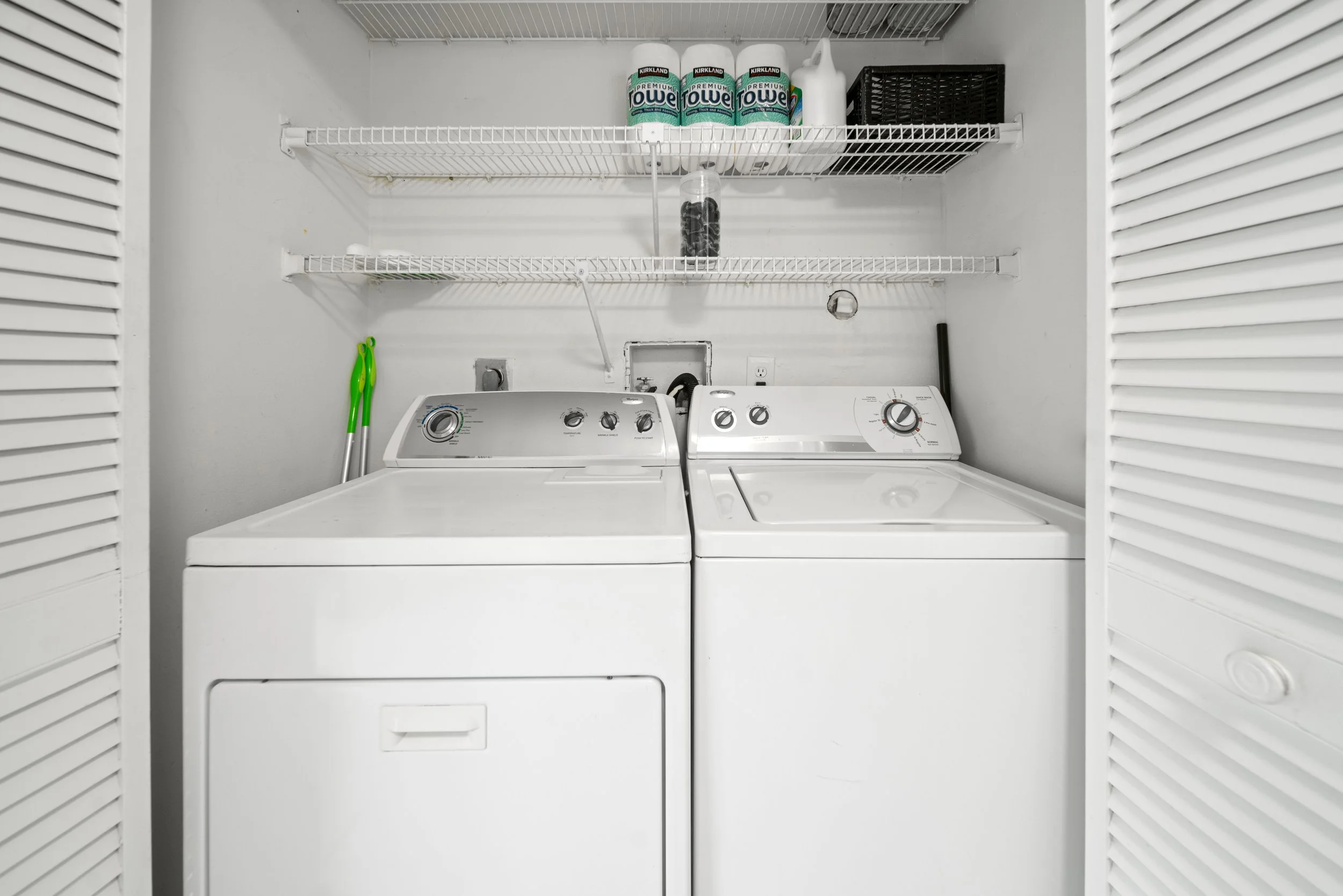 A laundry room with a white washer and dryer, wire shelving above with three Kirkland paper towel rolls, a black basket, and cleaning supplies. Two green handled mops are leaning against the wall on the left.