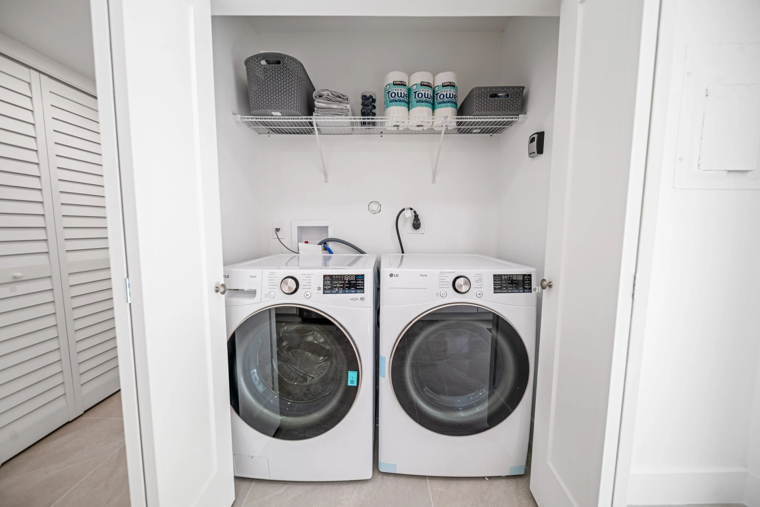A laundry closet with a white LG washer and dryer side by side, a wire shelf above holding paper towels, tissue boxes, and storage baskets, with a wall-mounted dryer vent and electrical outlets.