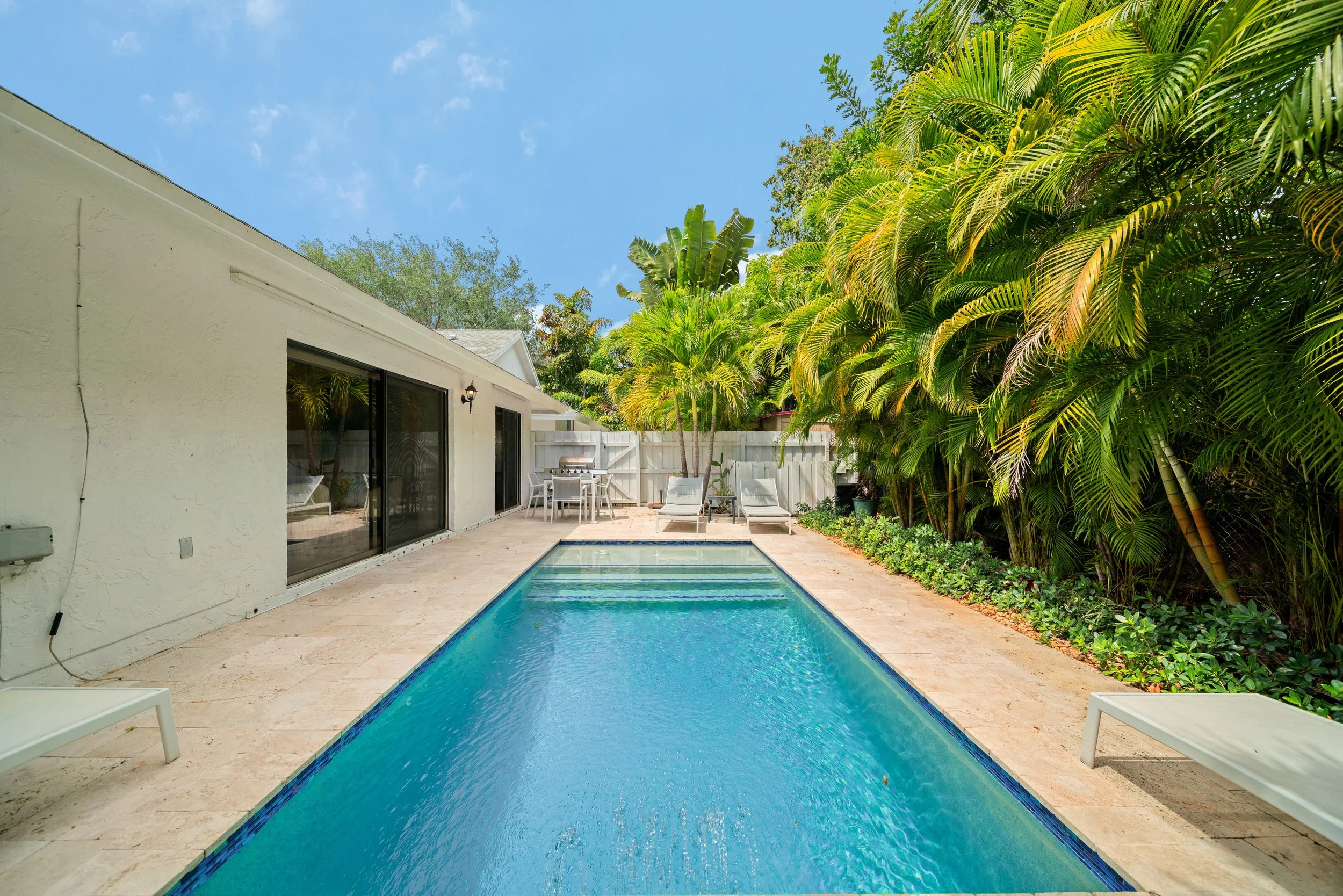 Backyard with swimming pool, white house wall with sliding glass doors, patio furniture including lounge chairs and table, surrounded by lush green tropical plants and trees, under a blue sky.