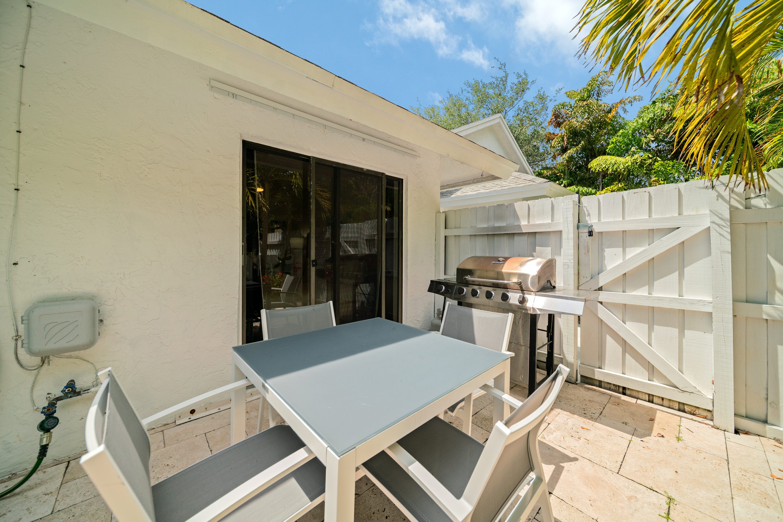 An outdoor patio area with a white table and six white chairs. There is a barbecue grill against a white wooden fence. The patio is paved with stone tiles, and there are leafy trees and a blue sky in the background.