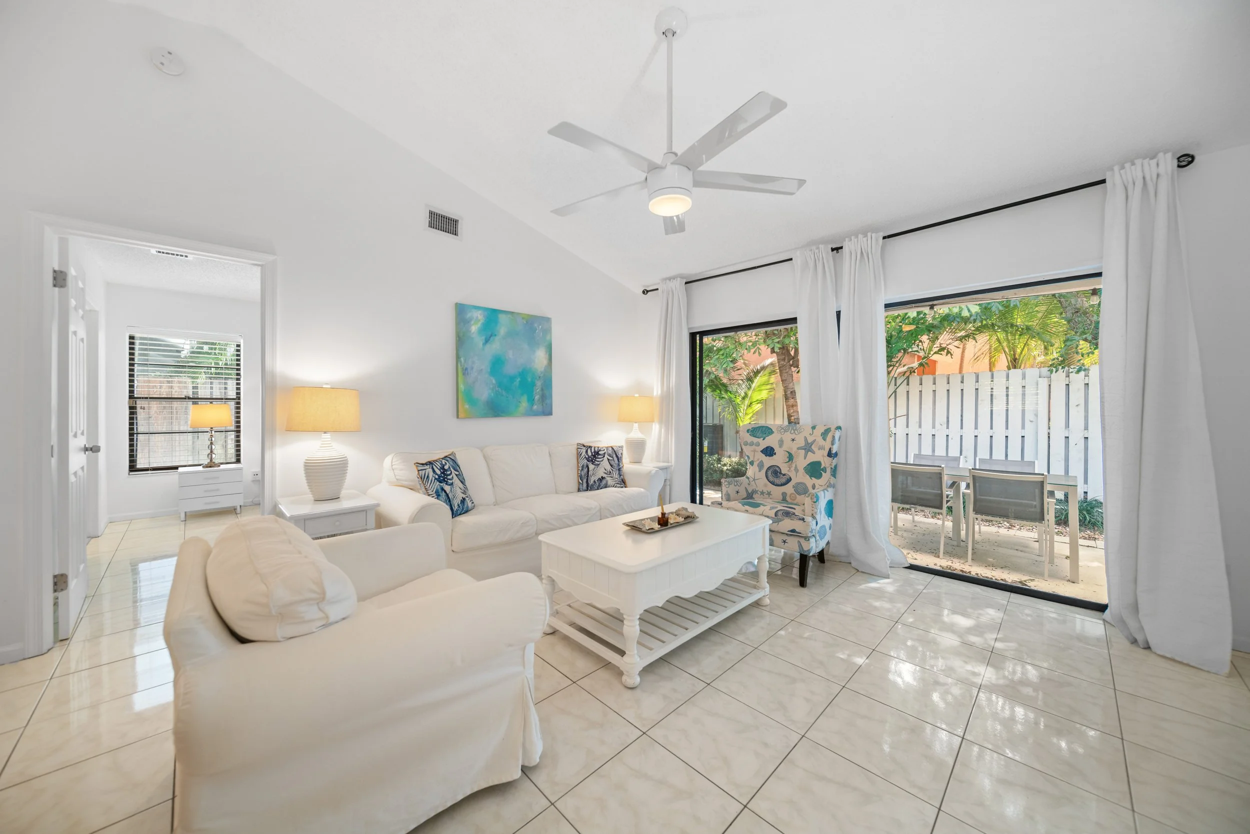 Living room with white furniture, a ceiling fan, and large sliding glass doors leading outside to a patio with tropical plants. White curtains and a colorful abstract painting on the wall.