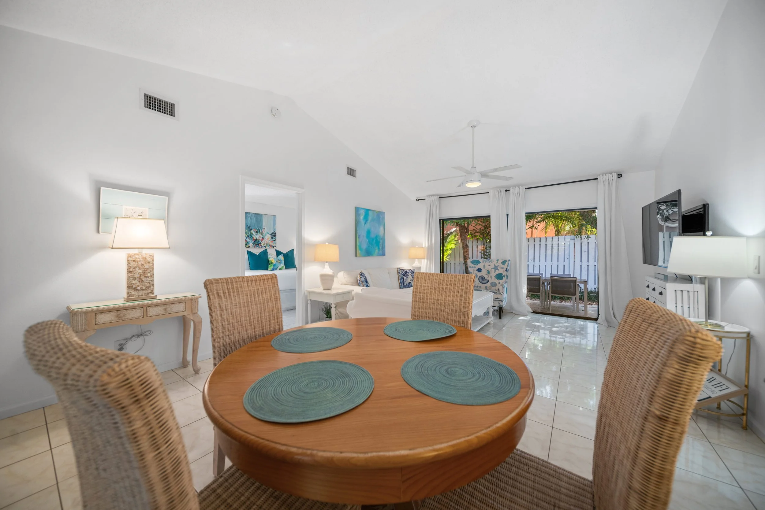 Living room with a dining table, wicker chairs, white sofas, colorful artwork, and large sliding glass door opening to an outdoor patio with plants.