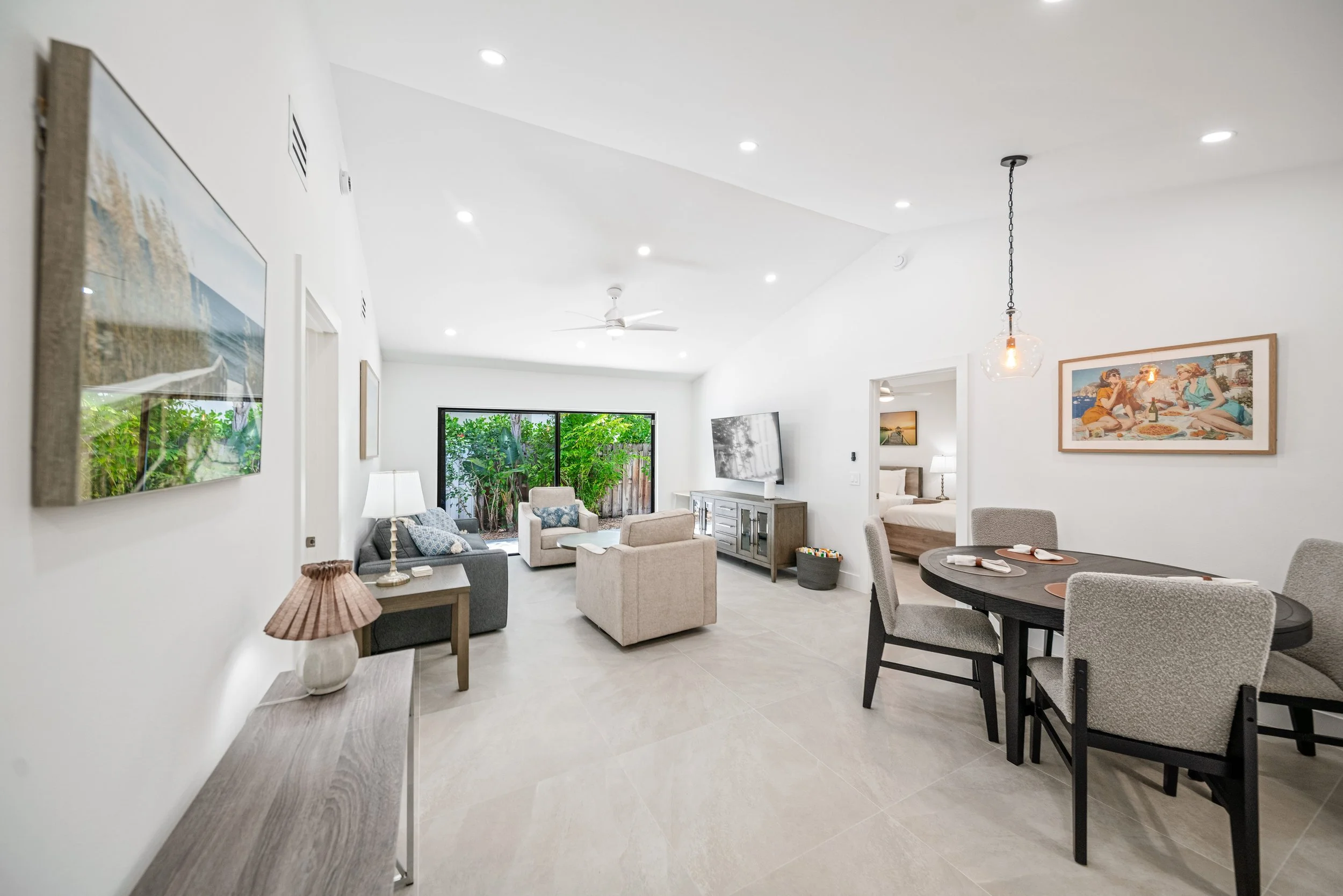 A bright and modern living room with white walls and ceiling, featuring a large sliding glass door opening to a garden, a ceiling fan, and recessed lighting. The room has a mix of neutral-toned furniture including armchairs, a dining table, and wall 