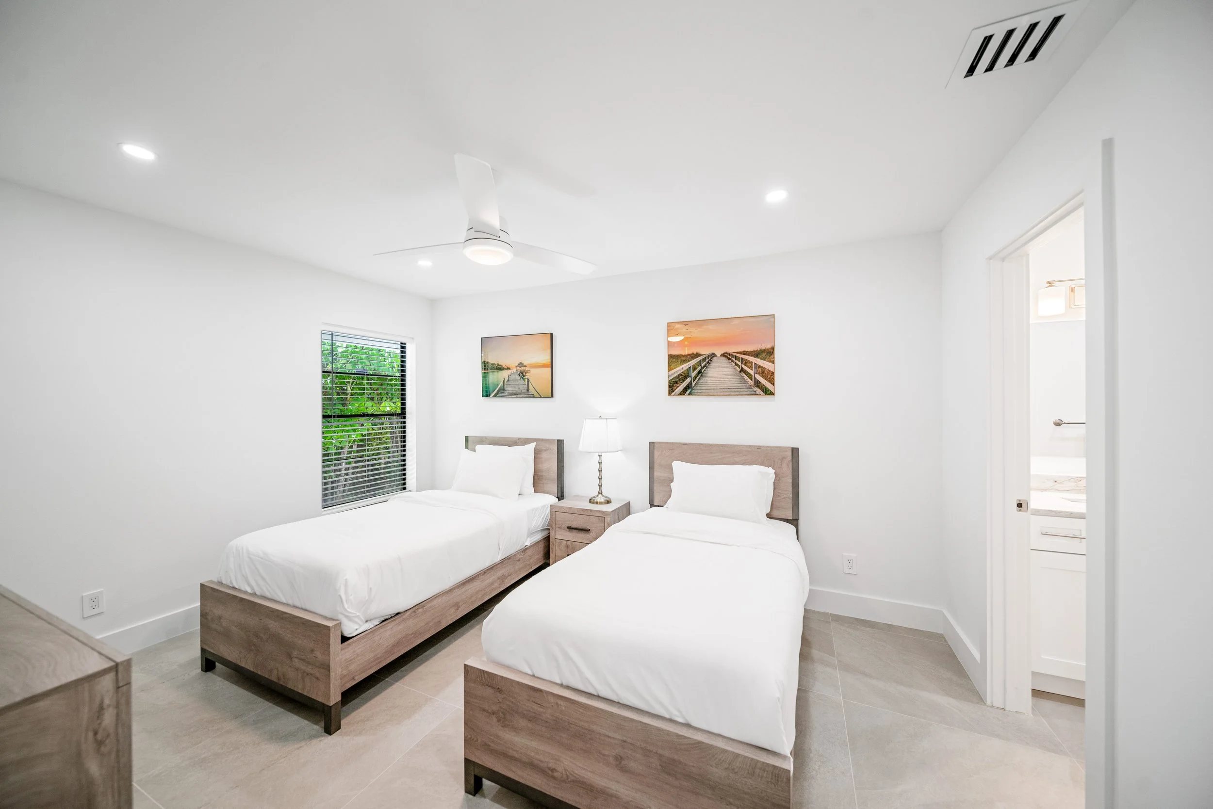 A bedroom with two twin beds with white bedding, wooden headboards, and nightstands. A lamp and framed pictures of a dock and sunset are on the wall. There is a window with blinds, a ceiling fan, and a doorway leading to a bathroom.