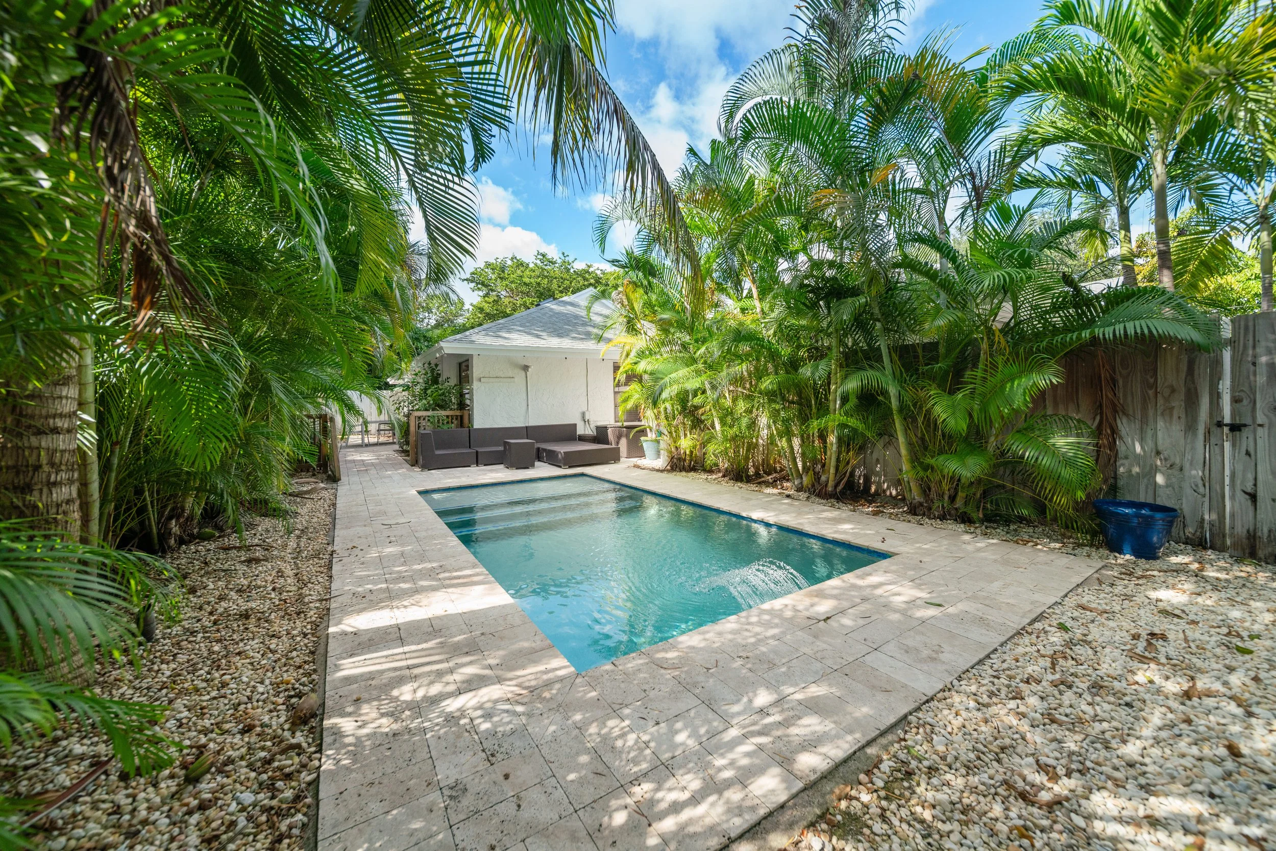 A backyard with a rectangular swimming pool surrounded by light-colored stone tiles and lush tropical palm trees. There is outdoor seating near the house and a blue planter on the right side.