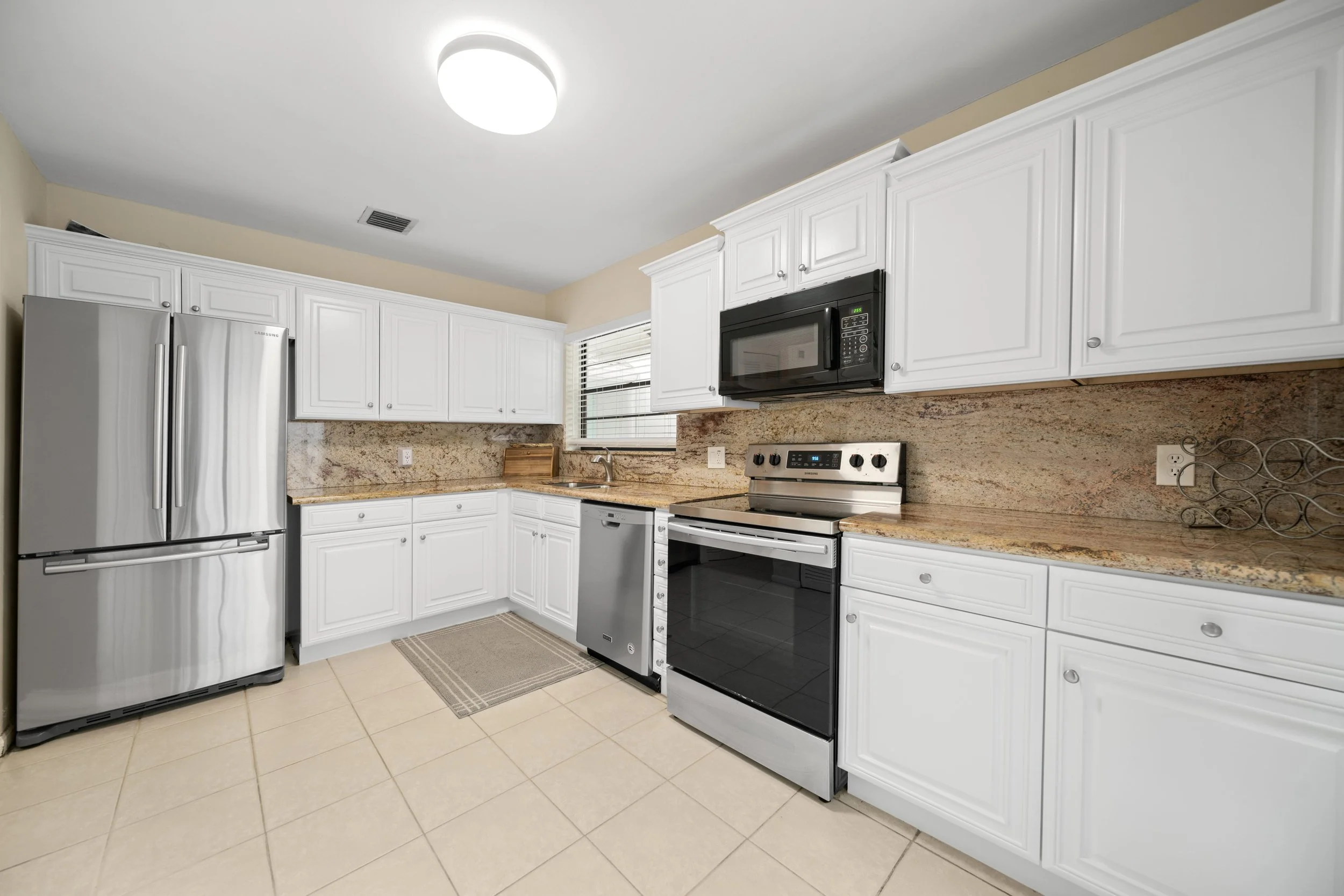 A clean kitchen with white cabinets, granite countertops, stainless steel appliances including a refrigerator, microwave, and oven, and a beige tile floor.