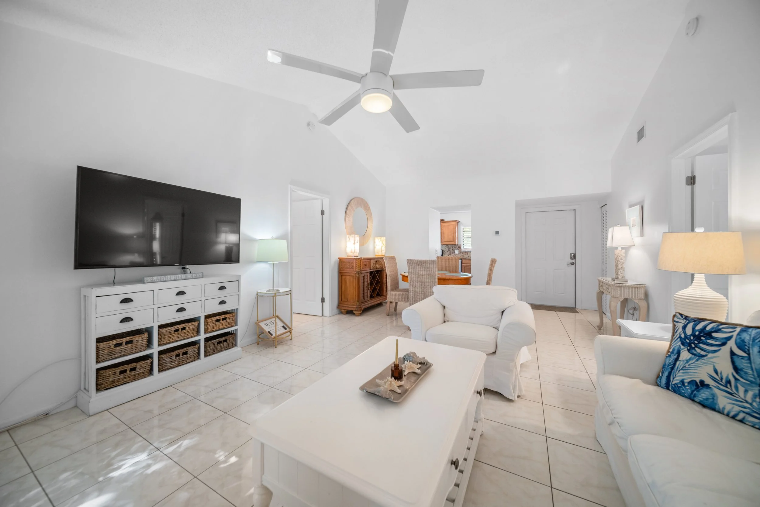 Bright living room with white walls and tile flooring, featuring a wall-mounted flat-screen TV, white media console with wicker baskets, seating with white upholstery and blue patterned pillow, wooden dining table with chairs, and a ceiling fan.