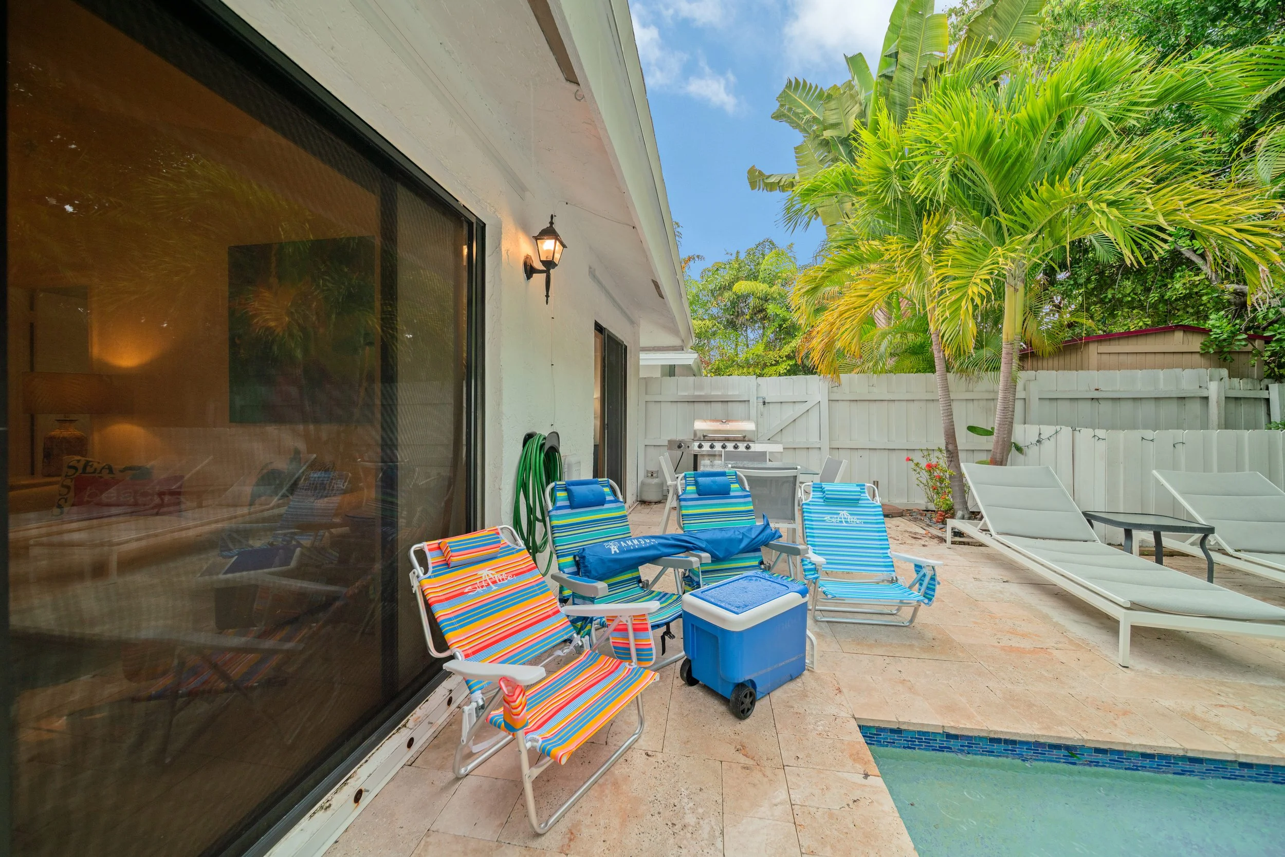 A backyard patio with multiple colorful lounge chairs, a grill, and tropical trees, with a view of a blue sky with some clouds.