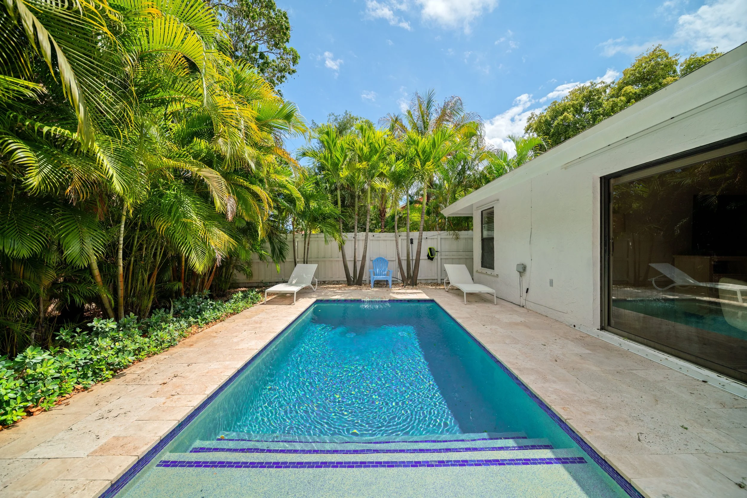 A backyard with a swimming pool, surrounded by green plants and trees, with a white house wall and sliding glass doors on the side.