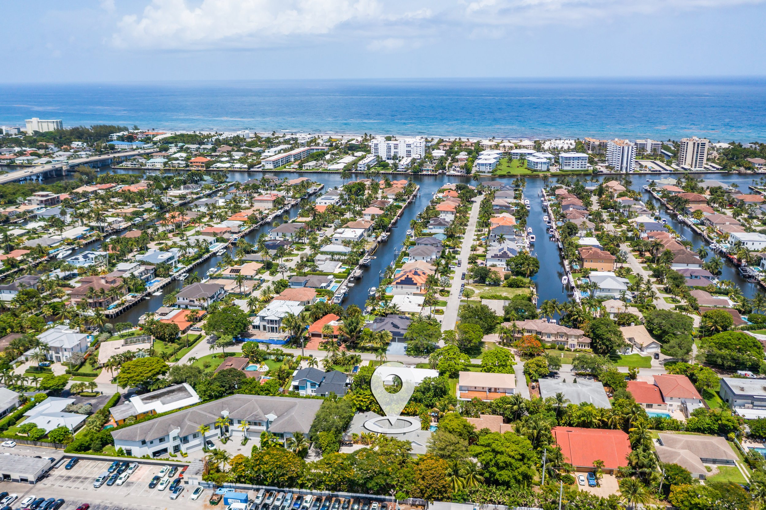 Aerial view of a coastal neighborhood with houses, canals, and palm trees near the ocean, with high-rise buildings in the background and an overcast sky.