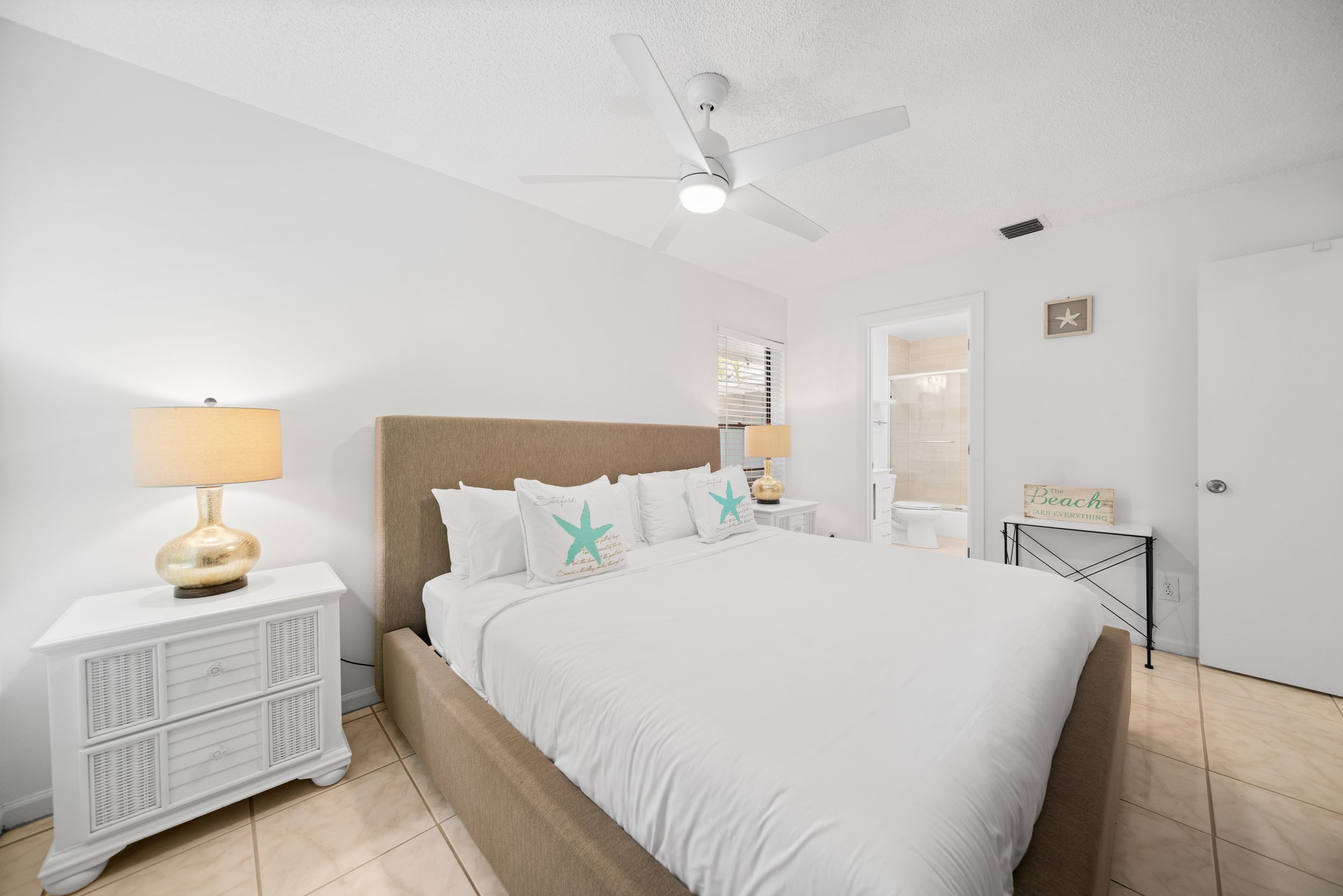 A bedroom with a beige upholstered bed, white bedsheets, and pillows with starfish prints. There are two white nightstands with lamps on either side of the bed, and a small table with a beach-themed sign near the doorway. The room has white walls, a 