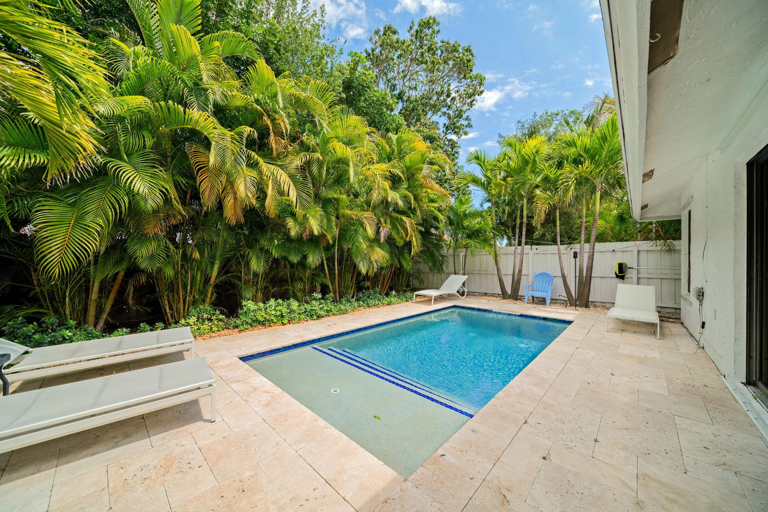 A backyard scene with a small swimming pool, surrounded by a patio with lounge chairs. Lush green palm trees and plants line the fence, and a white house wall with a window and some outdoor equipment are visible. The sky is partly cloudy with patches