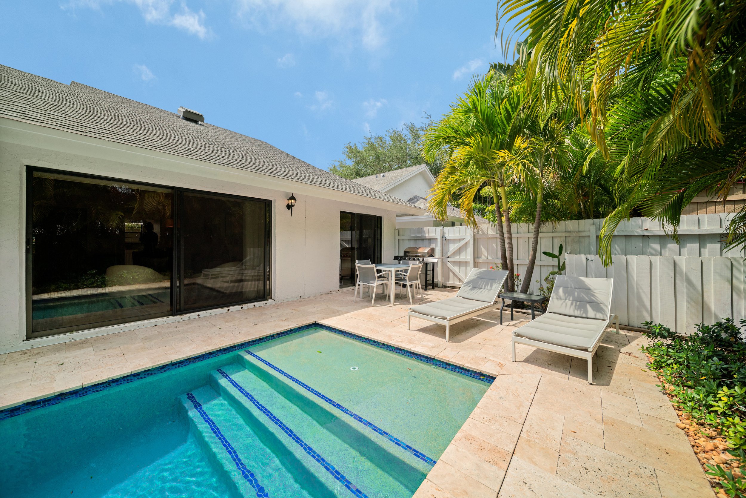 Backyard patio with a swimming pool, lounge chairs, a small table, a barbecue grill, tropical plants, and a house with sliding glass doors under a partly cloudy sky.