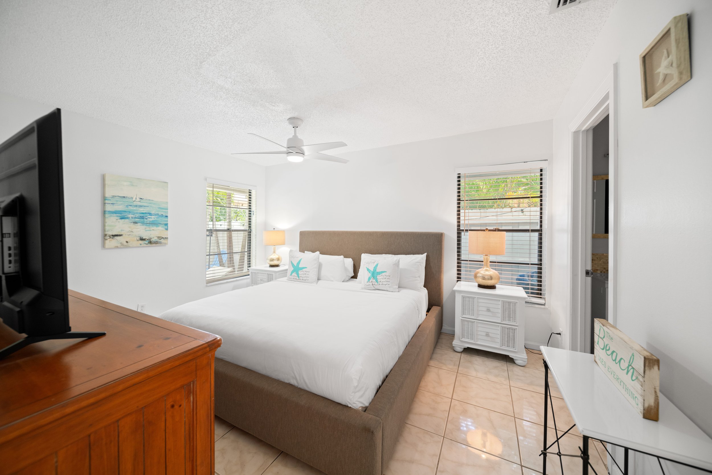 A bedroom with a beige bed, white bedding, and two starfish pillows. There are windows with white blinds, a nightstand with a lamp, and a ceiling fan. A painting of a beach scene hangs on the wall. A wooden dresser and a white table with a sign readi