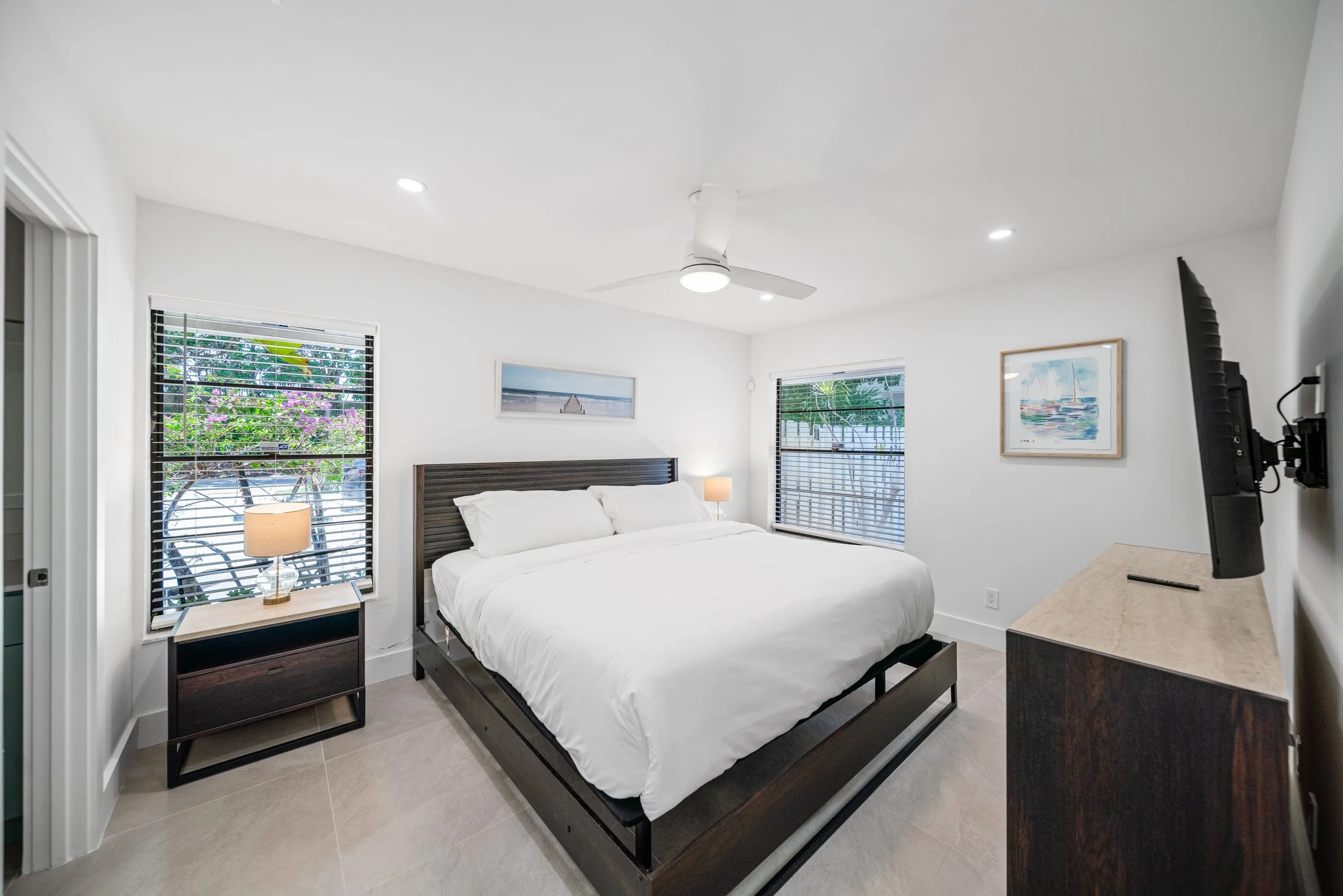 A modern bedroom with a white bed, wooden headboard, and two nightstands with lamps. The room has large windows with blinds, and artwork on the walls. There is a ceiling fan and a flat-screen TV mounted on the wall.