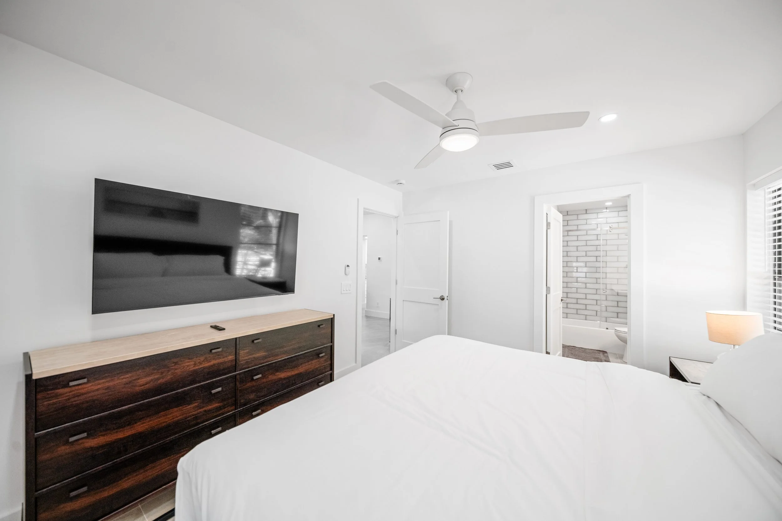 A bedroom with white walls, a ceiling fan, flat-screen TV, wooden dresser, white bed, and an open door leading to a bathroom with white subway tiles.