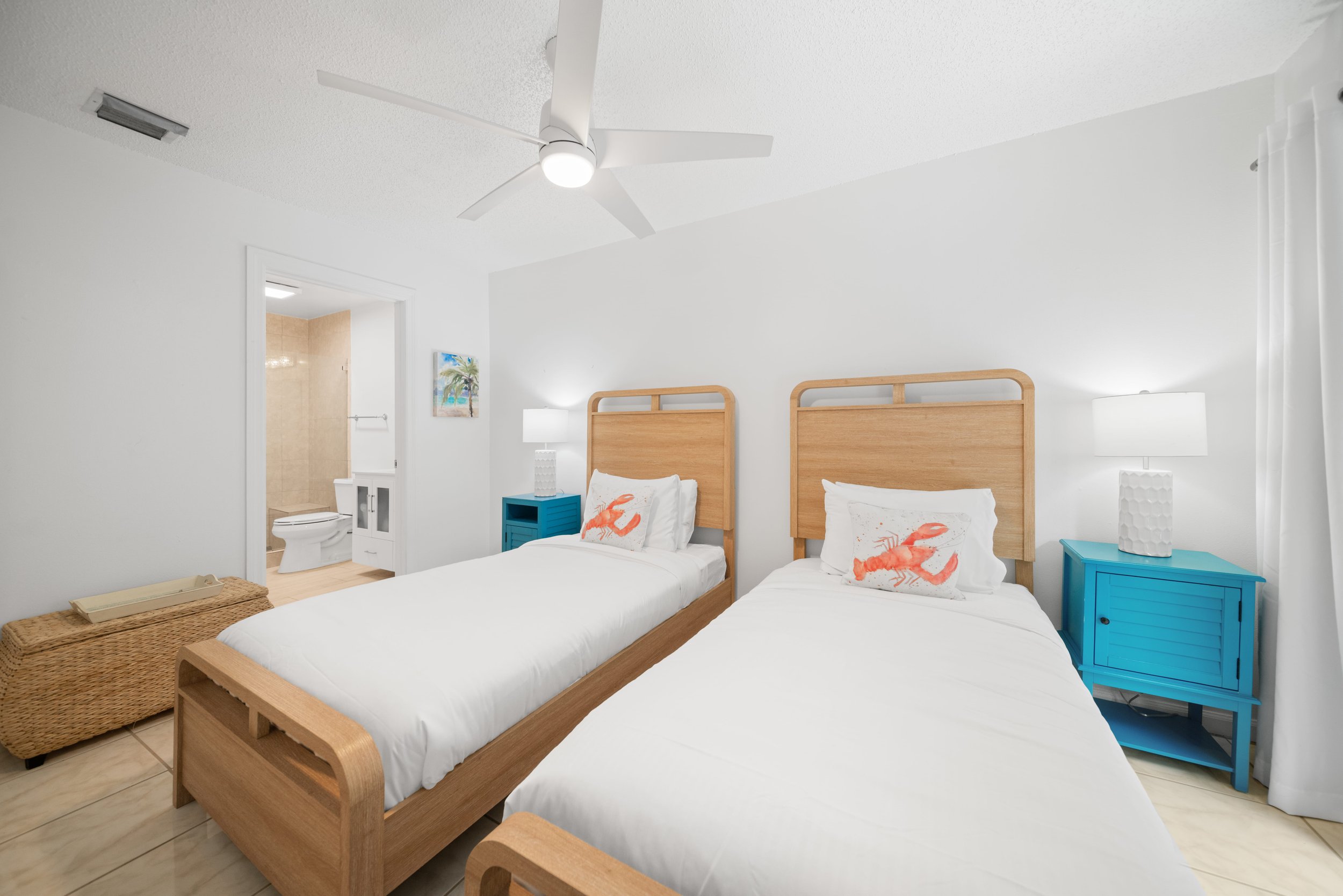 A motel room with two twin beds featuring lobster-themed pillows, a blue nightstand, and white lamps, with a doorway leading to a bathroom visible in the background.