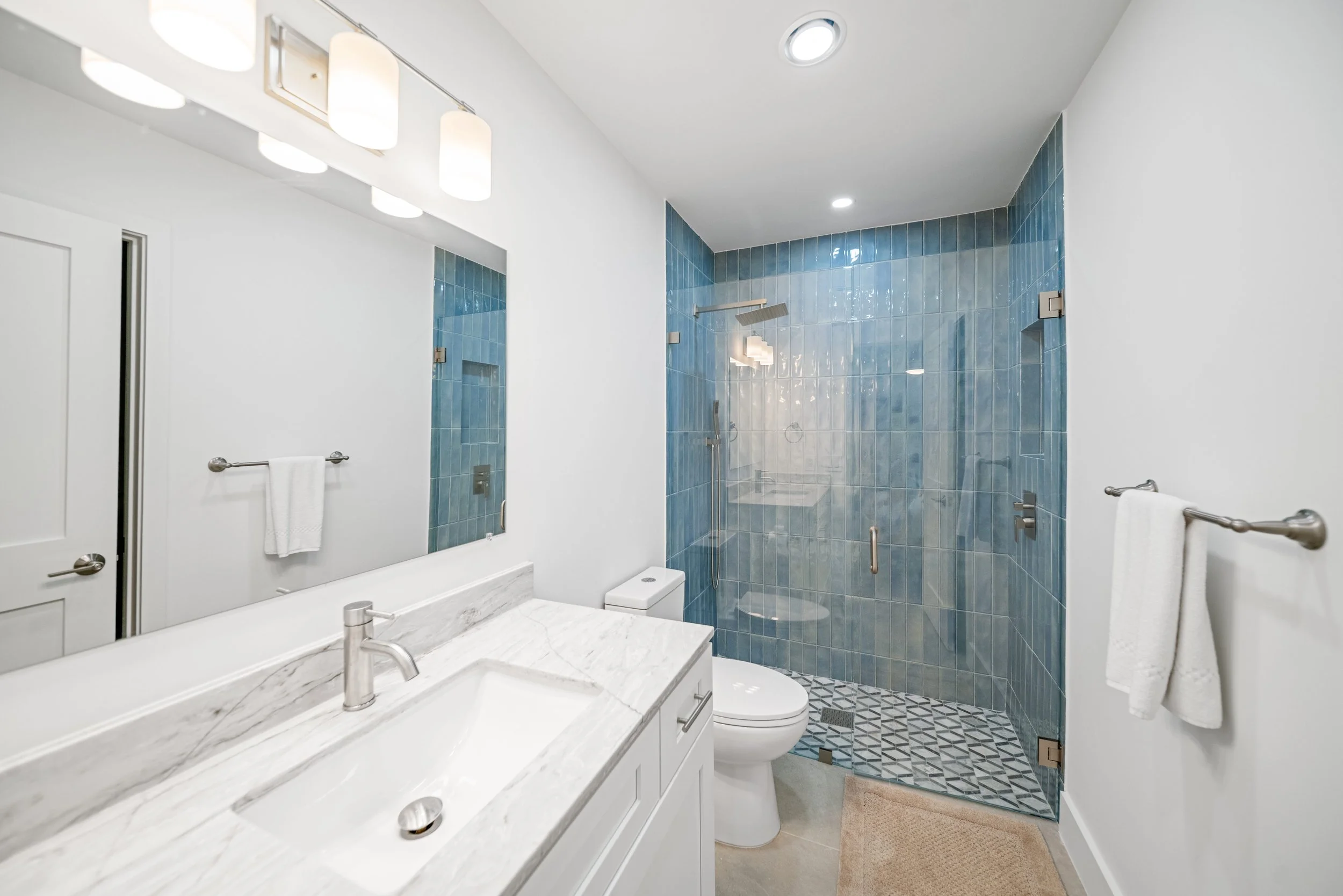A modern bathroom with a white marble countertop and sink, a wall-mounted mirror, a toilet, and a clear glass shower with blue tiles and a rainfall showerhead.