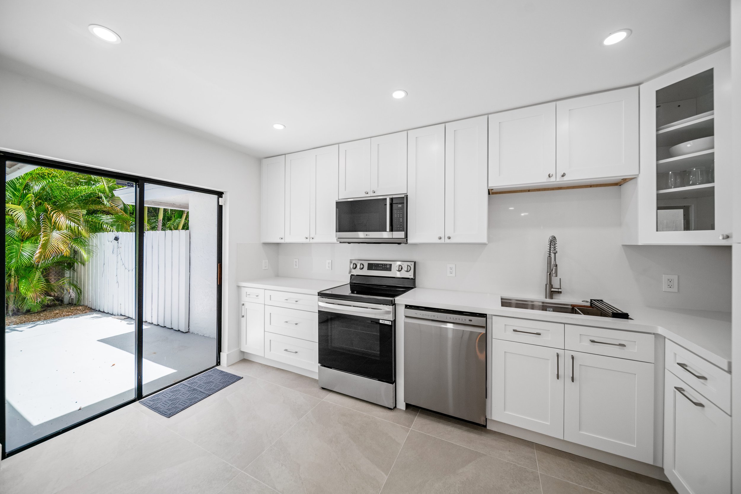 Modern white kitchen with stainless steel appliances, white cabinets, and a sliding glass door leading outside to greenery.