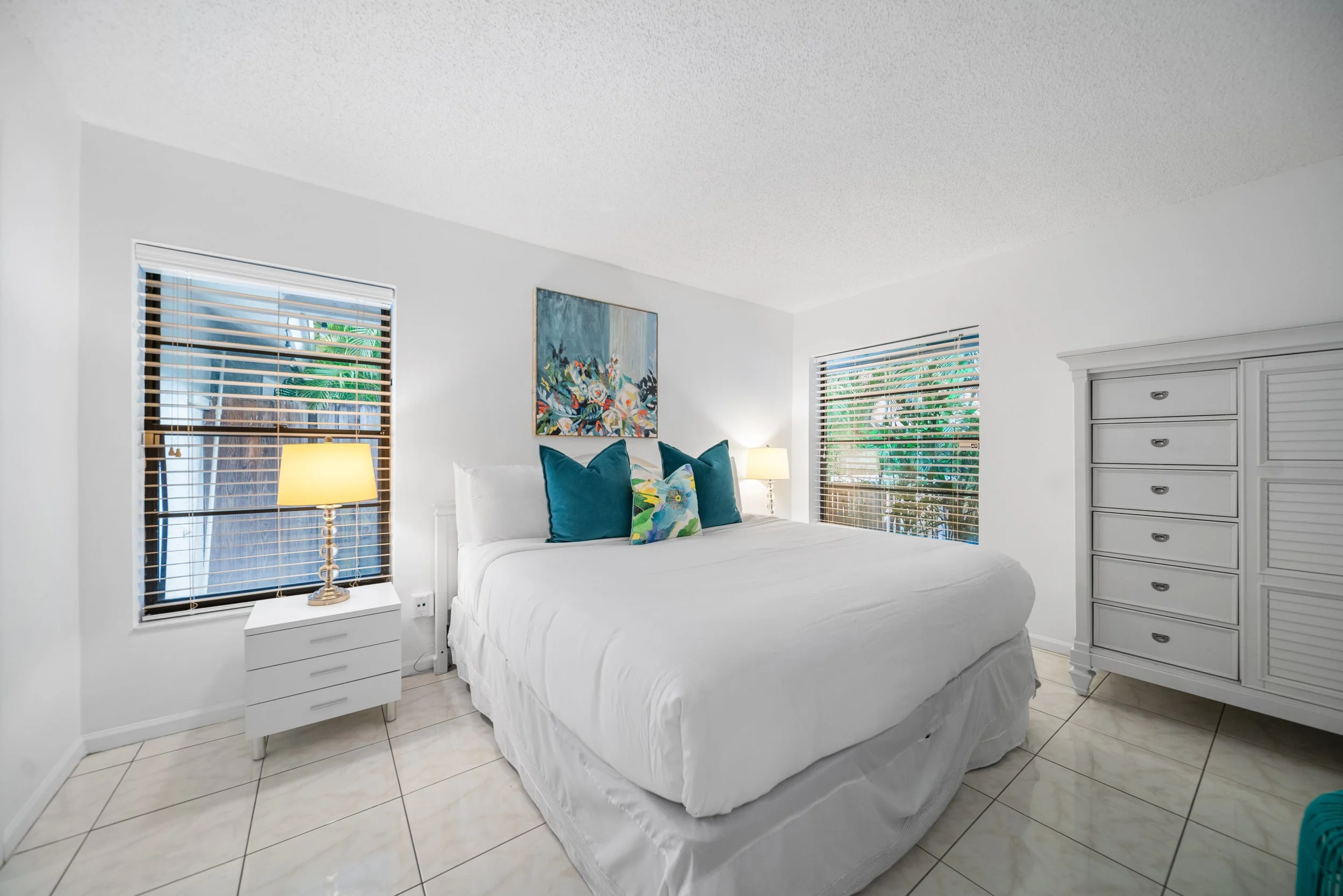 A bright bedroom with white walls, a white bed with colorful throw pillows, two side tables with lamps, and a tall white dresser. Two windows with wooden blinds let in natural light. There is a blue and white floral artwork on the wall.
