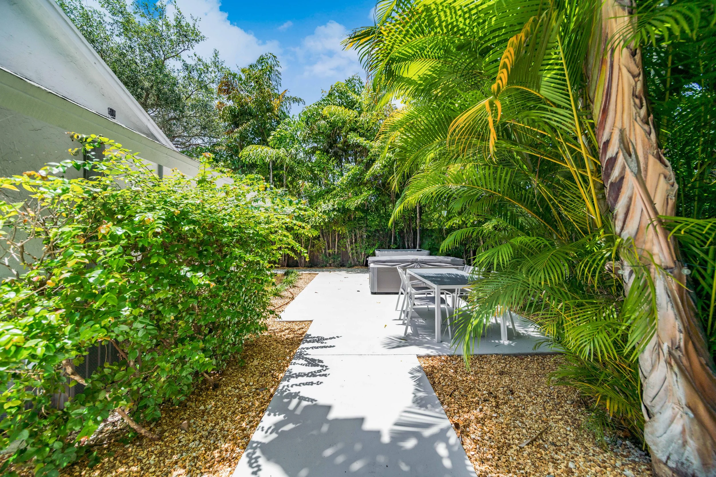 A backyard patio with a concrete path, outdoor furniture, surrounded by lush green plants and trees, with a partly cloudy sky overhead.