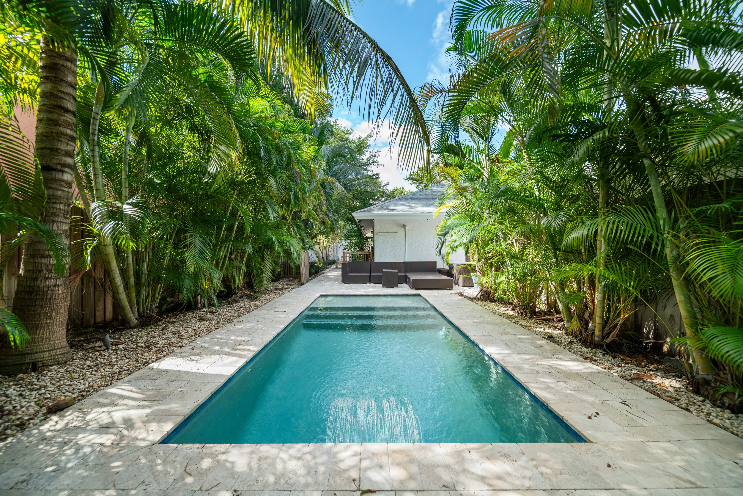 A backyard scene with a rectangular swimming pool surrounded by lush tropical plants and palm trees. There is a white house with a gray roof in the background, and outdoor seating with cushioned furniture visible near the house.