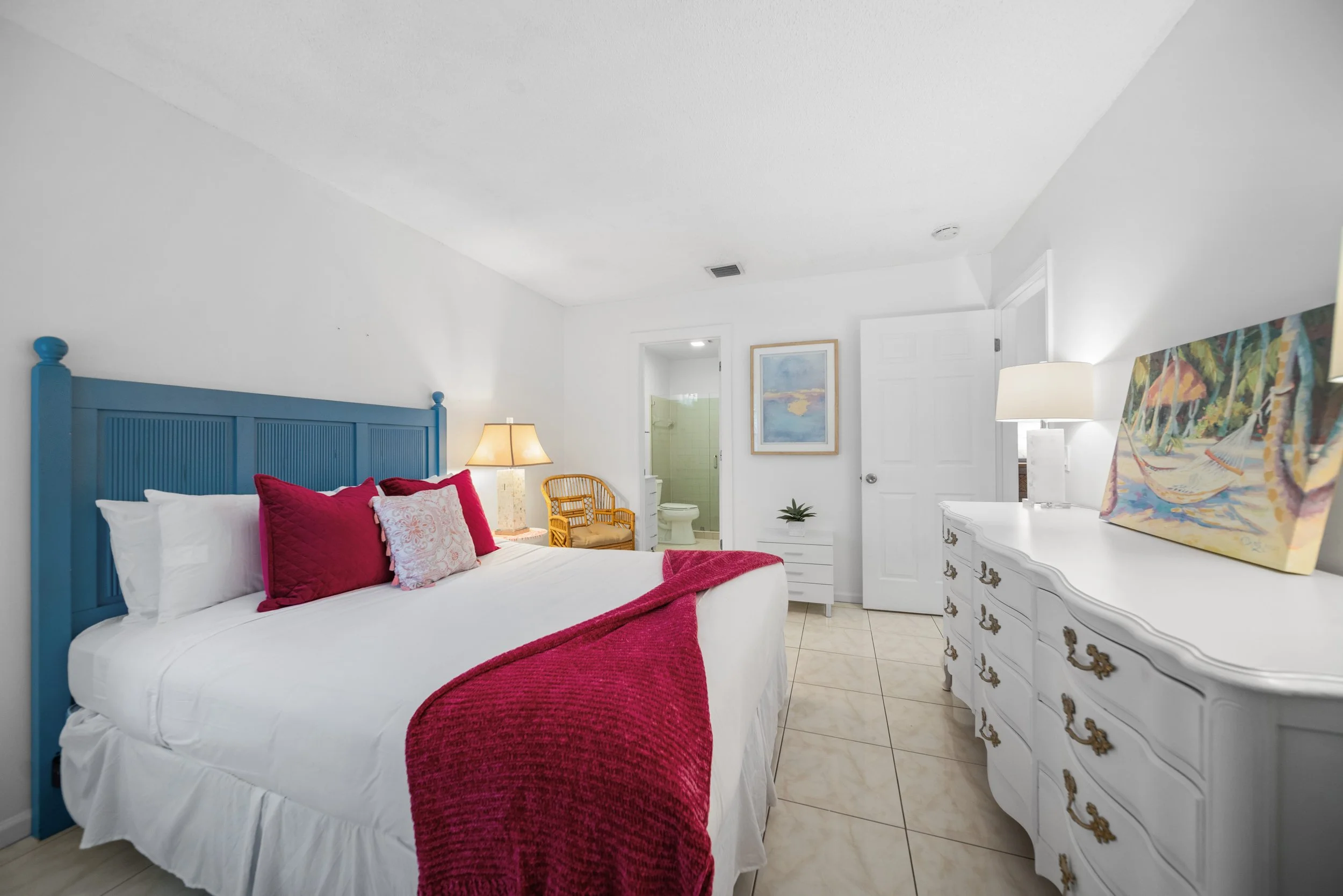 A bedroom with a bed that has a blue headboard, white bedding, and red and white pillows. There is a nightstand with a lamp, a rattan chair, and a dresser with a colorful painting. The room has white walls, tiled floor, and a visible bathroom in the 