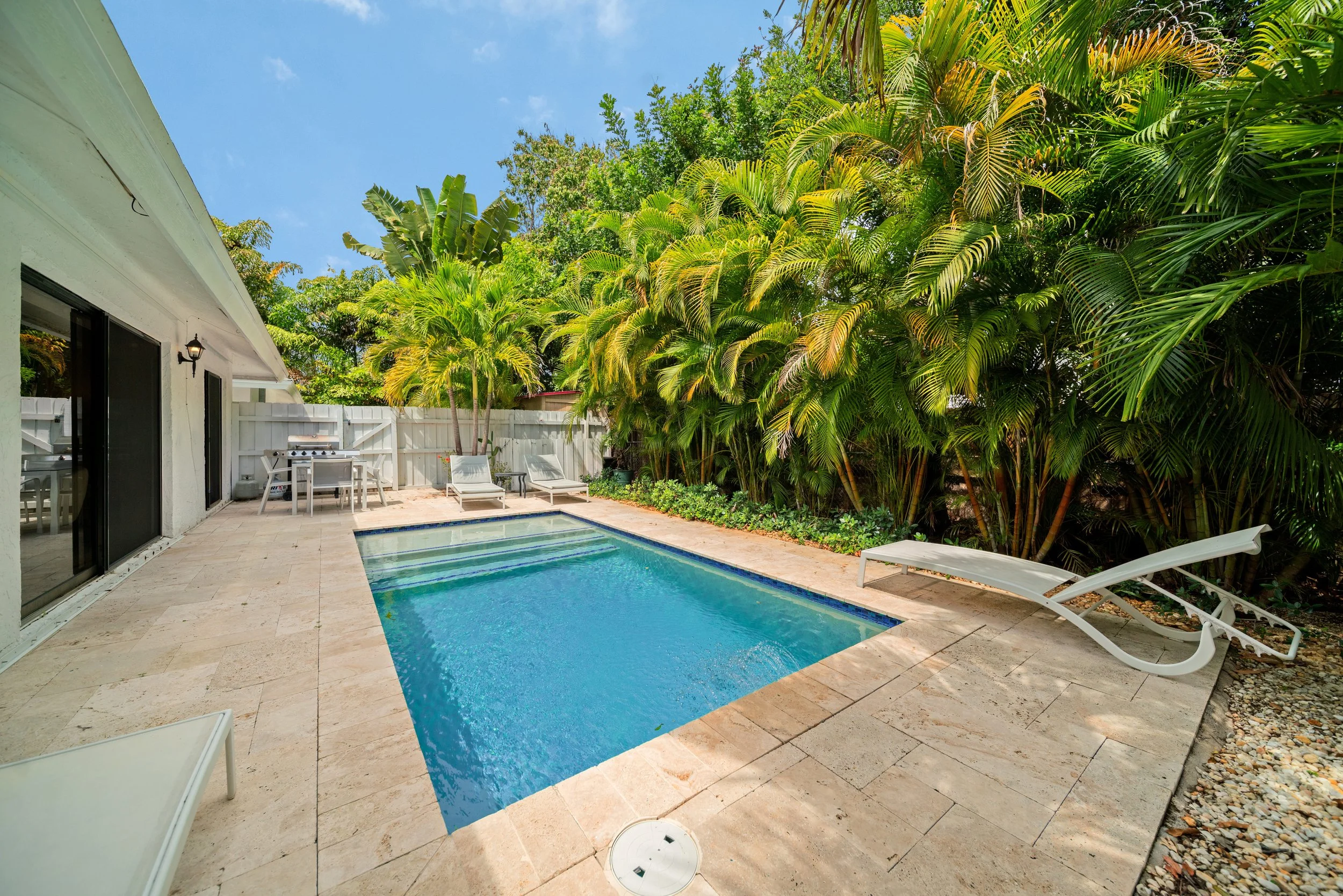 A backyard with a swimming pool, surrounded by a white fence, lush green trees, and lounge chairs. Part of a house with sliding glass doors and an outdoor light fixture is visible.