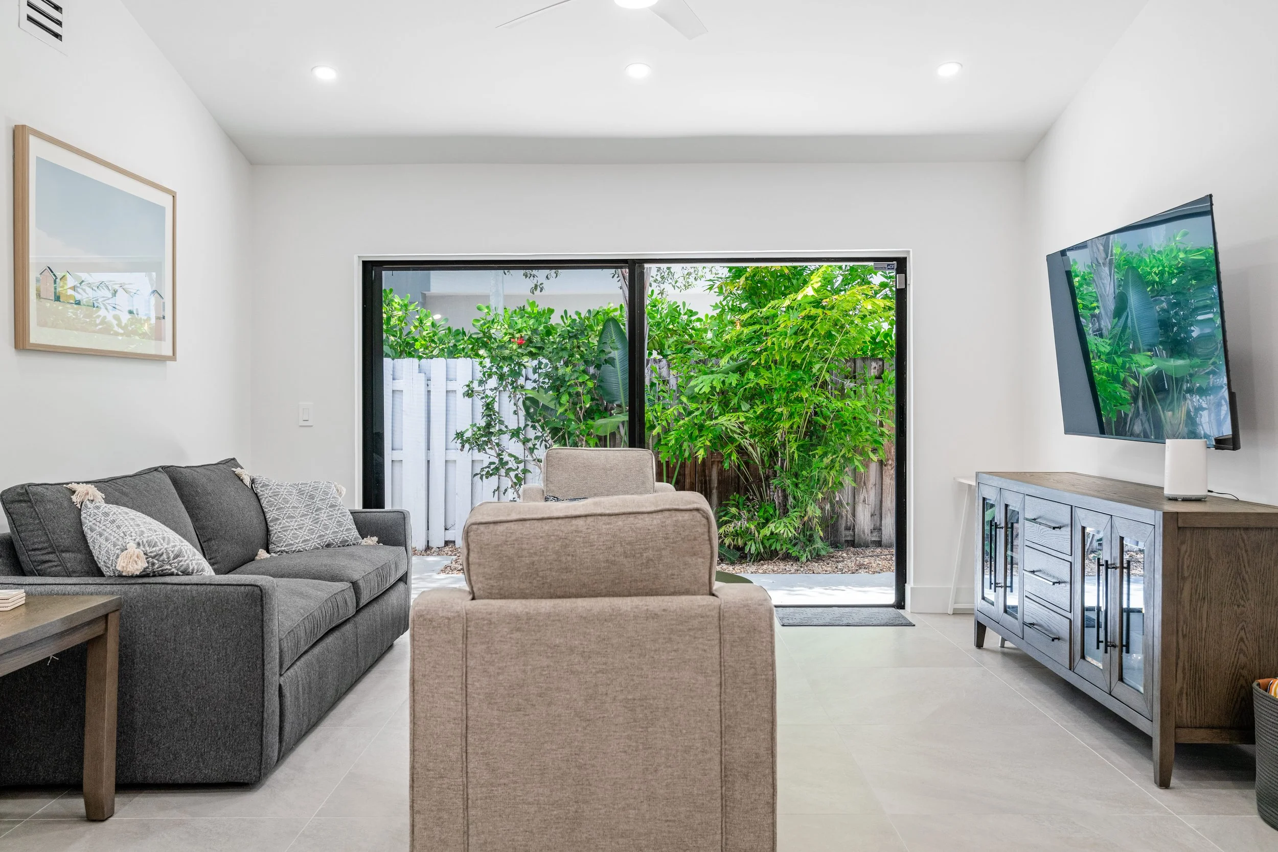 Living room with gray sofa, beige armchair, and wooden side table, large sliding glass door opening to a garden with lush green foliage, TV mounted on the wall, and modern white ceiling with recessed lighting.