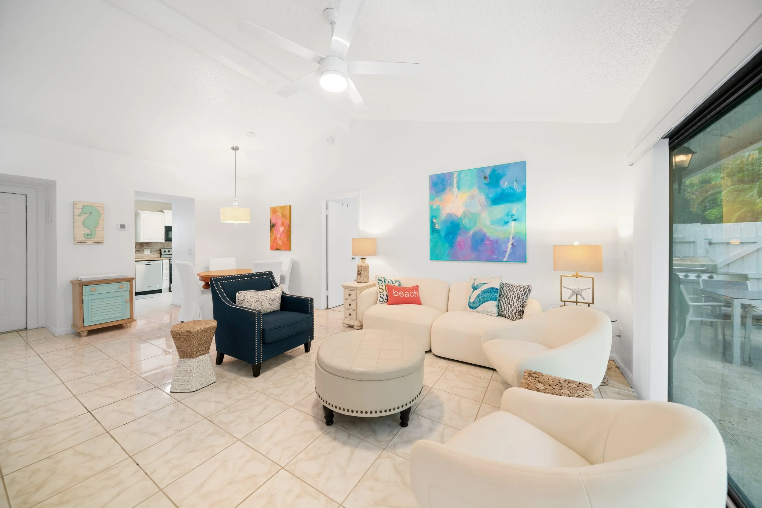 Living room with white walls and tiled floor, featuring a white ceiling fan, colorful artwork, a beige sofa with decorative pillows, a blue armchair, a lamp, and a sliding glass door leading outside.
