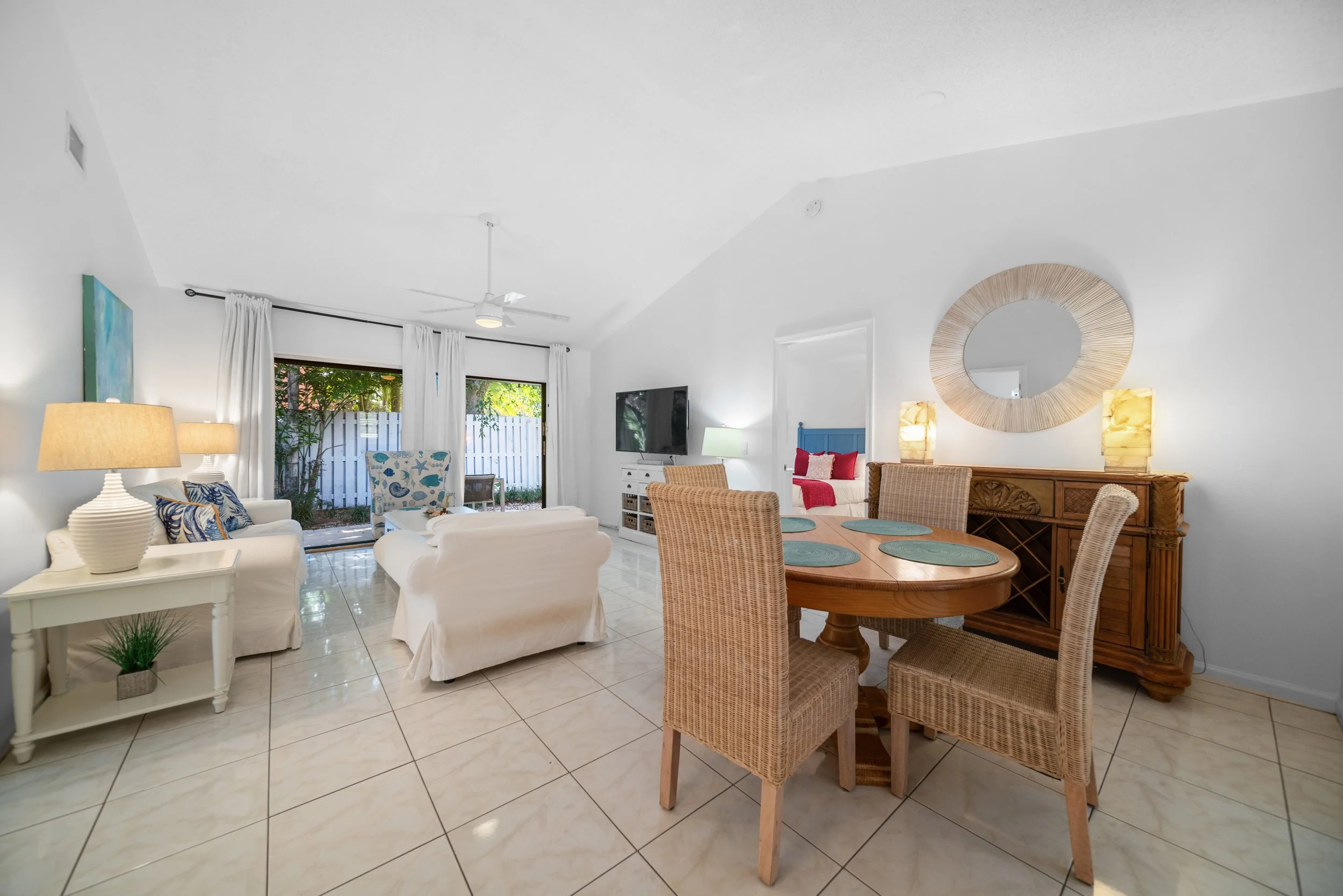Living room with white sofas, wicker chairs, a round wooden dining table with plates, a sideboard, a wall mirror, a TV, and sliding glass doors leading outside to greenery.
