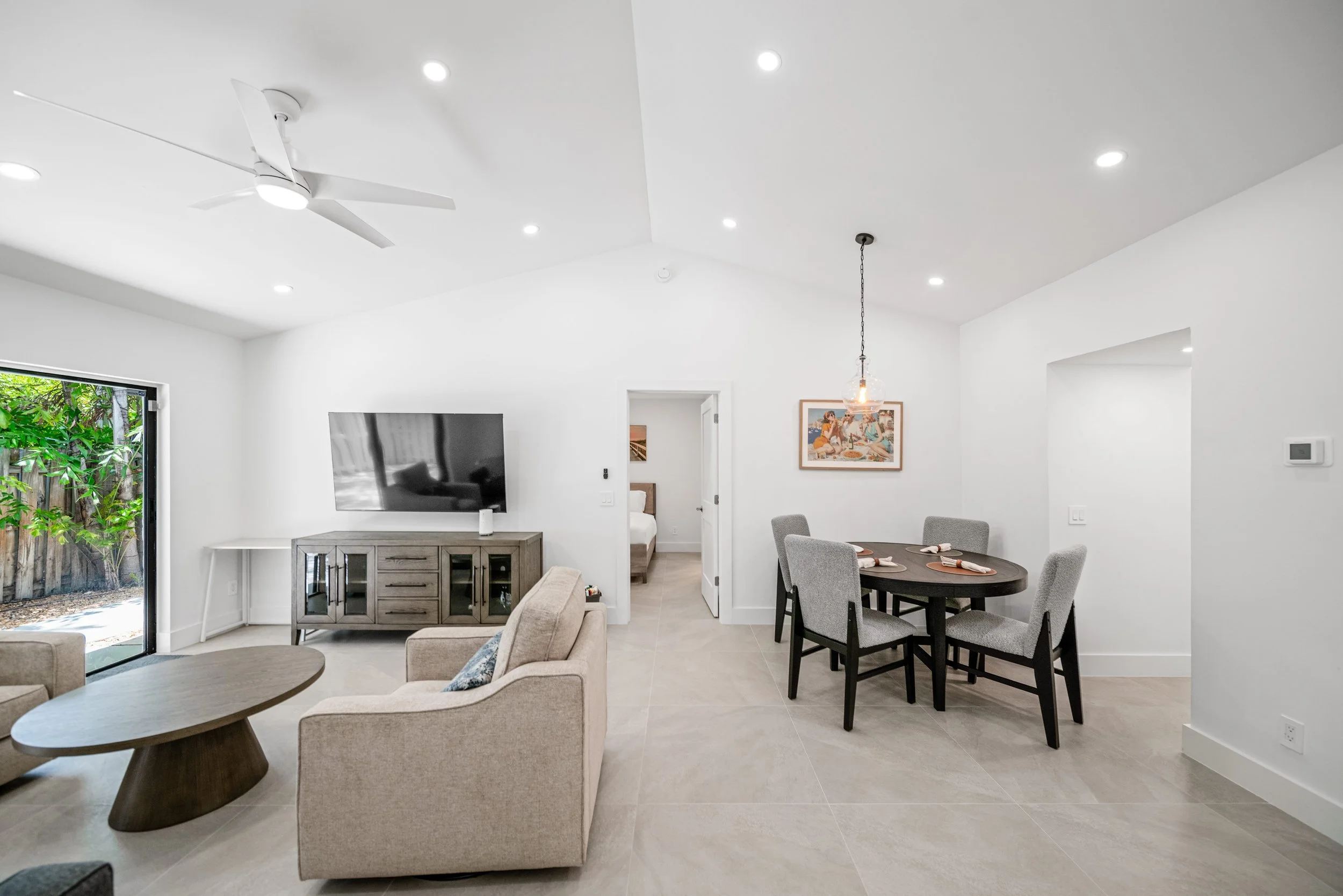 Open plan living and dining area with white walls, neutral furniture, a round wooden coffee table, a flat-screen TV on a wooden console, a dining table with six gray chairs, a painting on the wall, a ceiling fan, and a sliding glass door leading outs