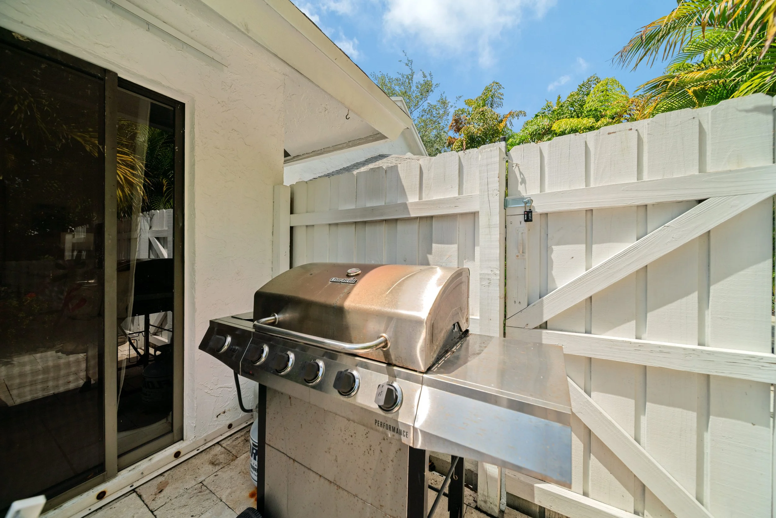 A backyard patio with a stainless steel gas grill, a white wooden fence, and lush green trees under a blue sky with some clouds.