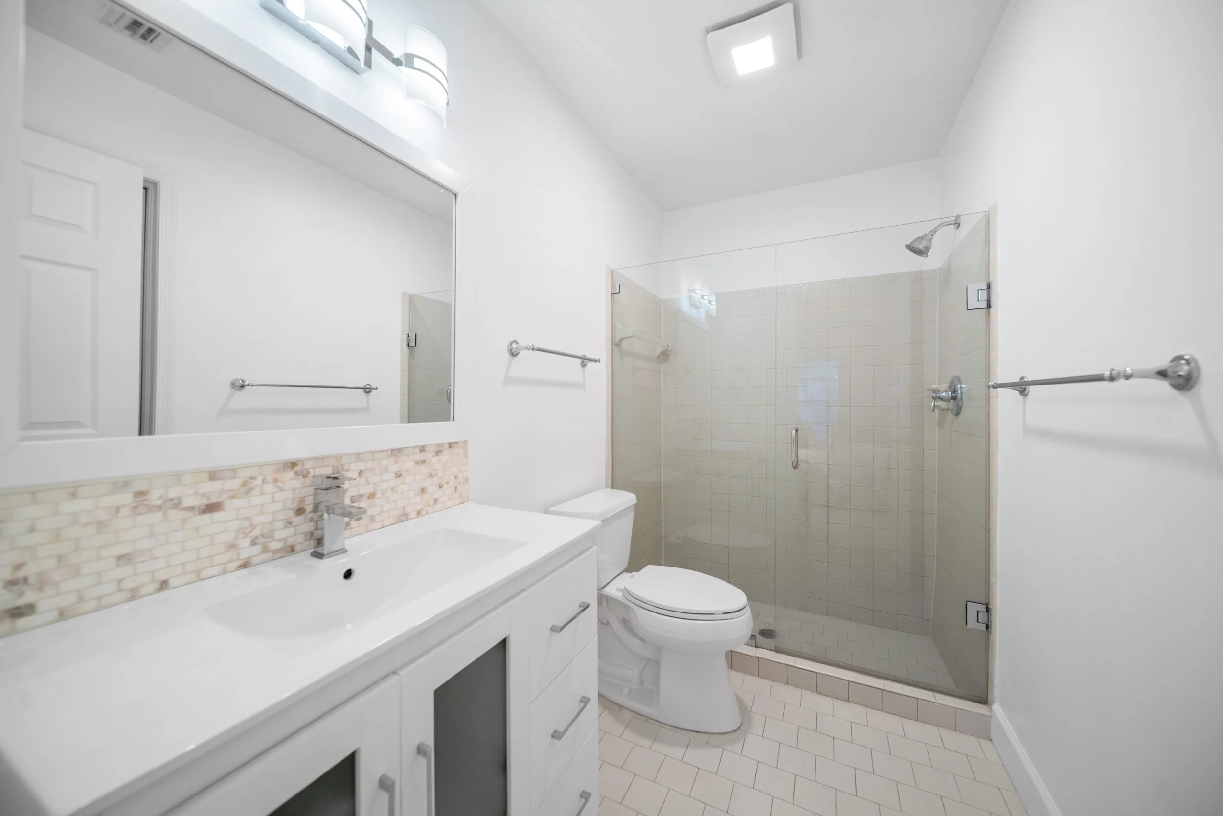A modern bathroom featuring a white vanity with a single sink, a large mirror, a toilet, and a walk-in glass shower with a sliding door. The bathroom walls are painted white, and the floor is tiled with small, light-colored tiles.