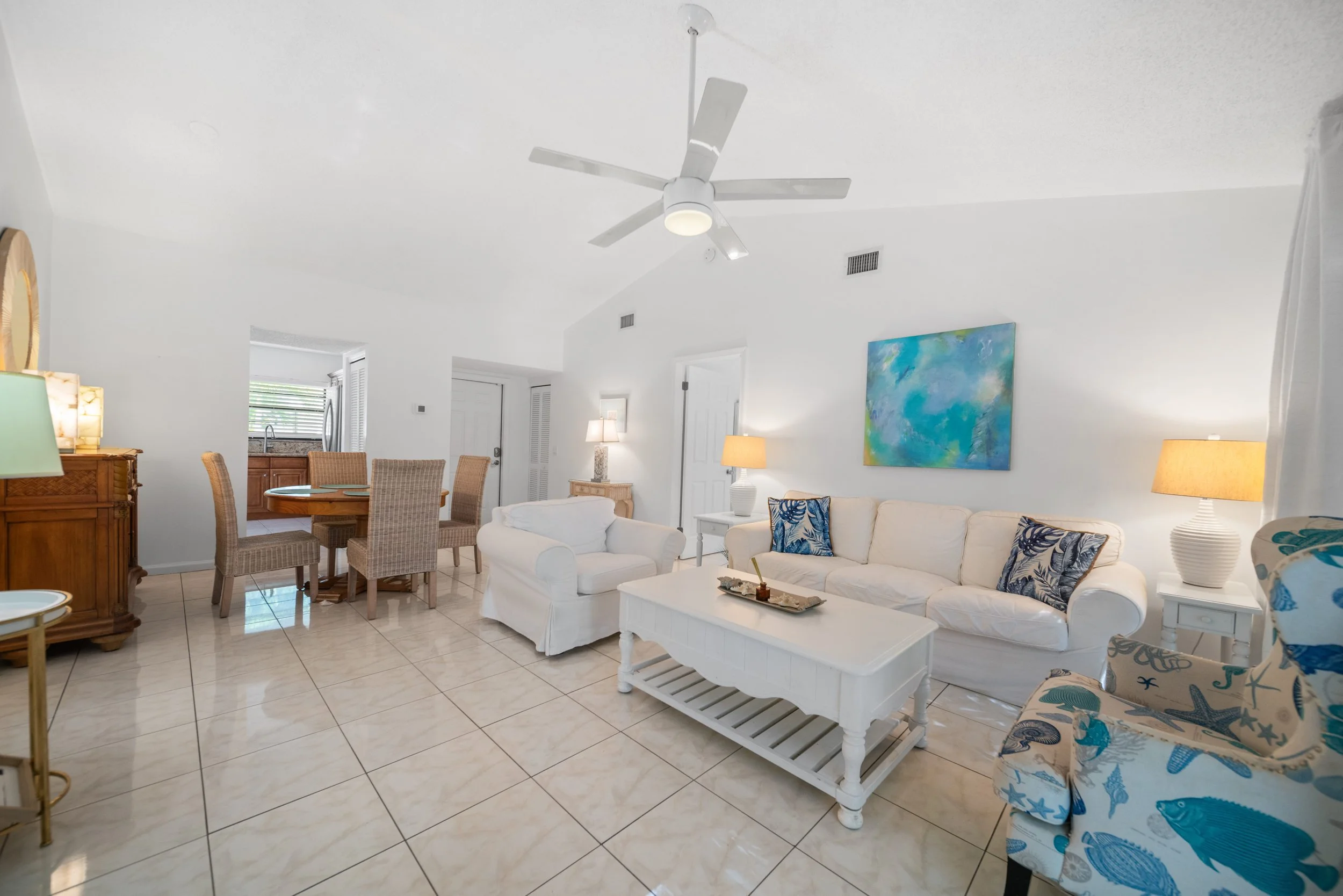 Living room with white walls, tiled floor, white sofa with blue and white patterned pillows, wooden dining table with four wicker chairs, kitchen counter with a stainless steel refrigerator, colorful abstract painting on the wall, and various lamps.