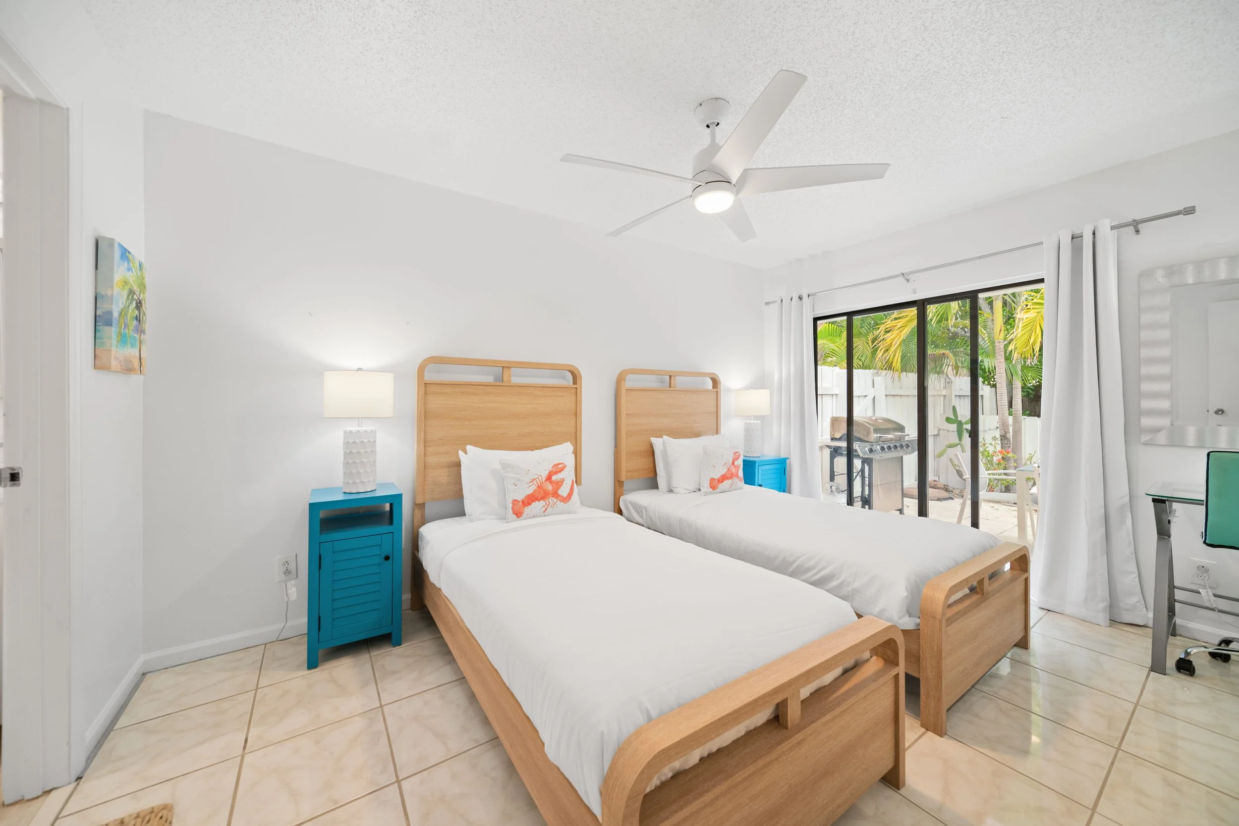 A bright bedroom with two twin beds made of light wood, each with white bedding and lobster pillows. A blue nightstand is between the beds with a white lamp, and a large window or sliding door with white curtains shows palm trees outside.