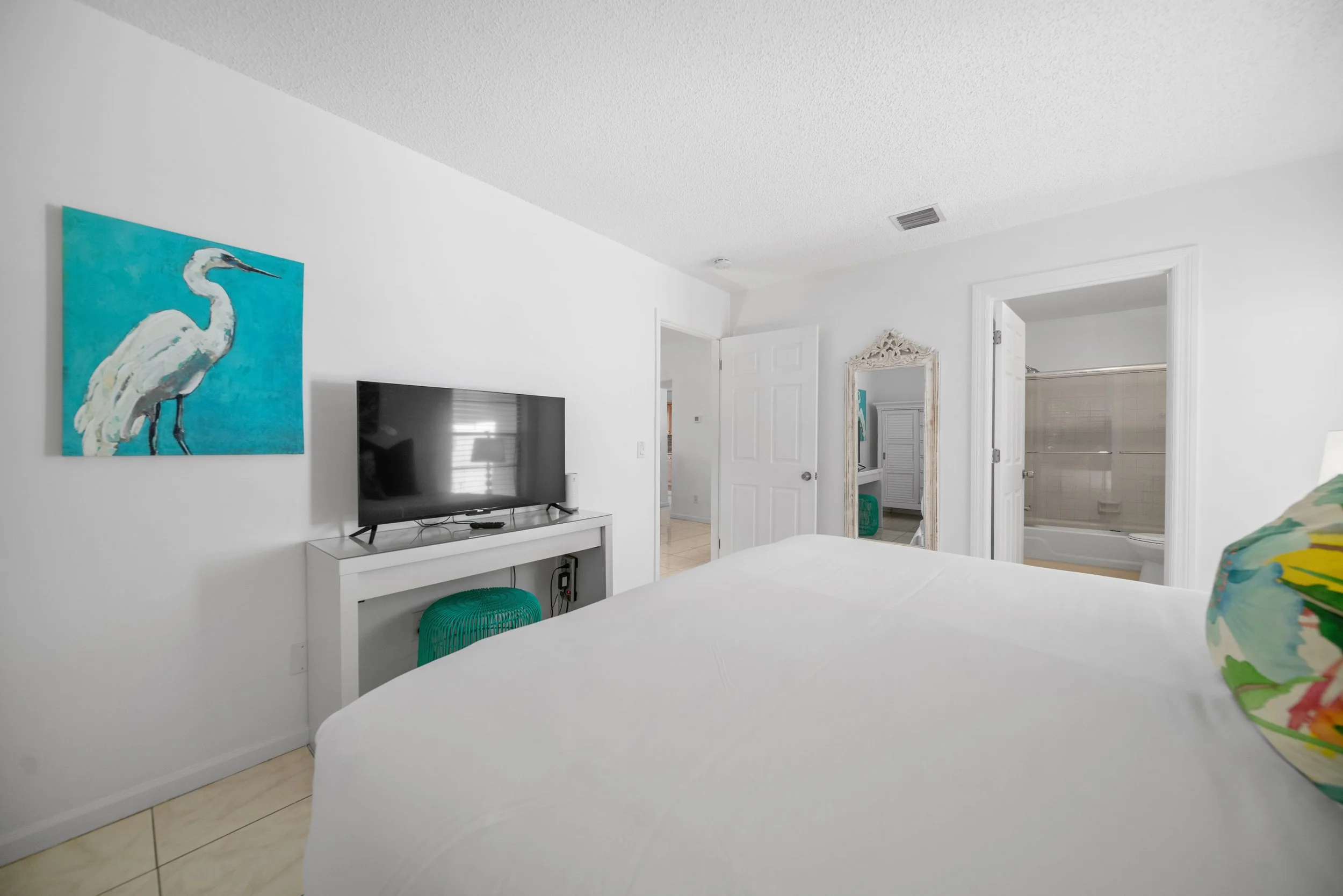 A bedroom with white walls and ceiling, a bed with white bedding, a flat-screen TV on a white desk, a colorful painting of a white heron on the wall, a small teal stool, a mirror with an ornate white frame, and an open door leading to a bathroom with