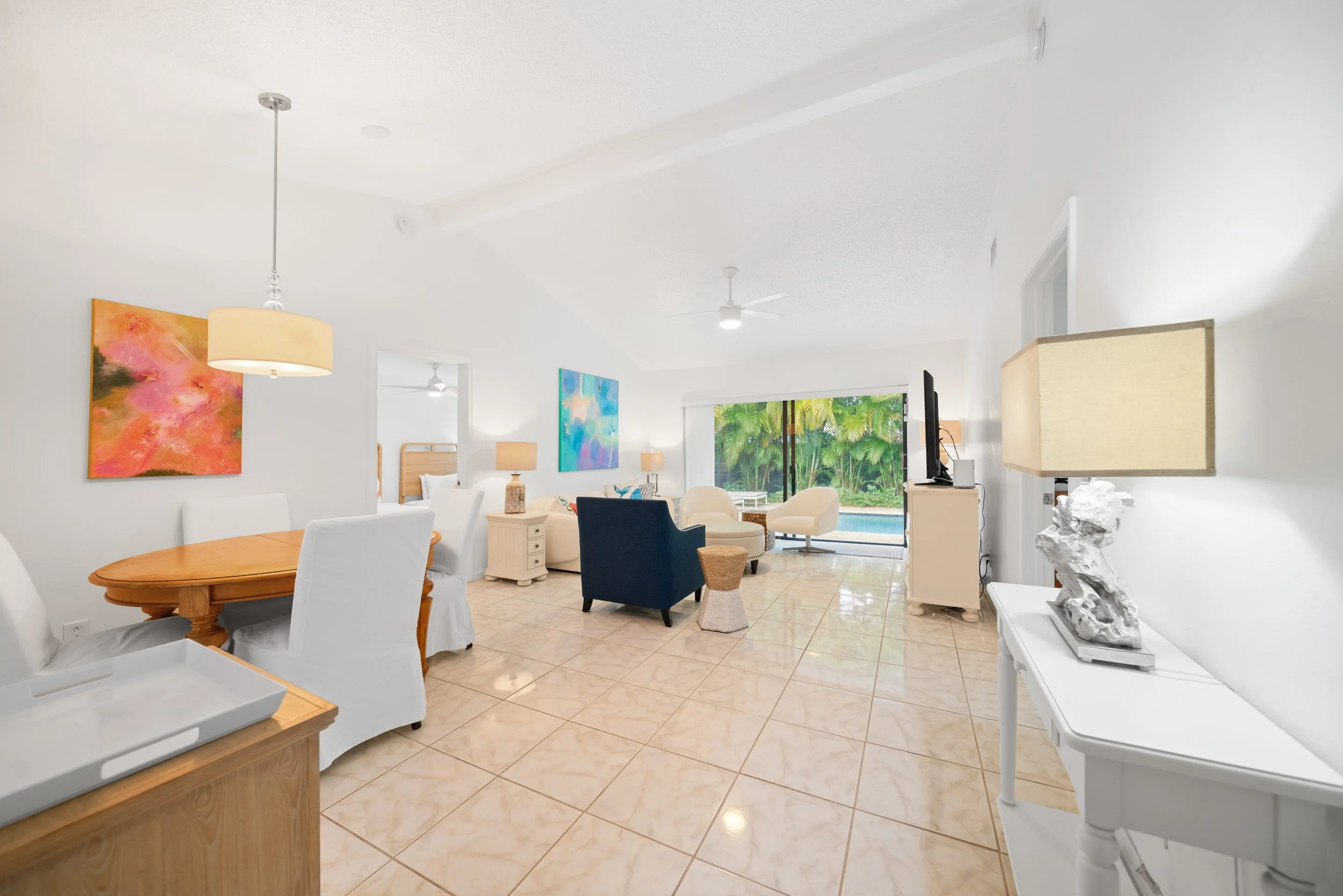 Bright, modern living room with white walls, tile flooring, colorful abstract paintings, white and dark blue armchairs, a round wooden dining table with white chairs, and a sliding glass door showing a lush green garden.