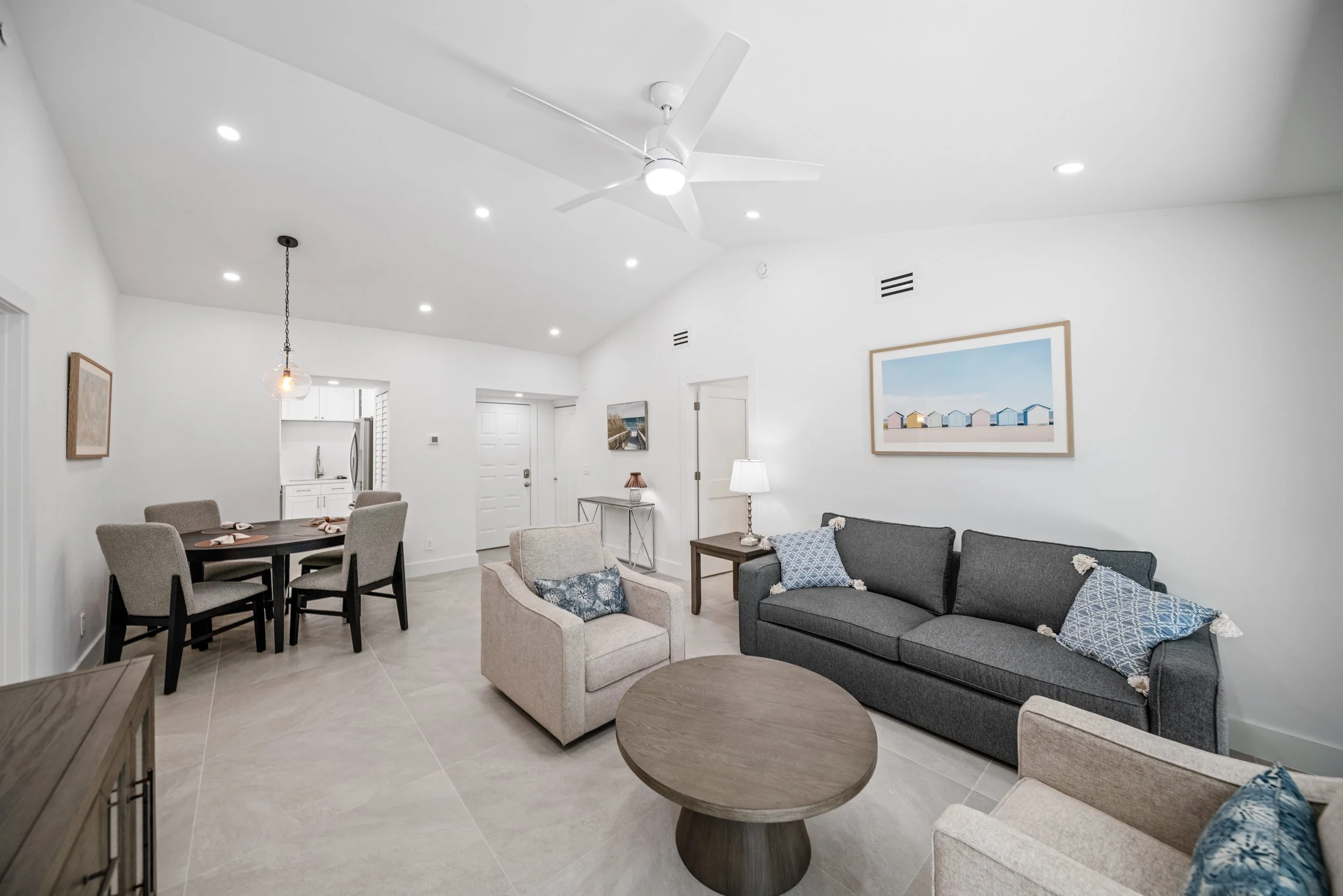 Living room with beige armchair, dark gray sofa with blue pillows, round wooden coffee table, white walls, framed art, ceiling fan, recessed lighting, and an open kitchen in the background.