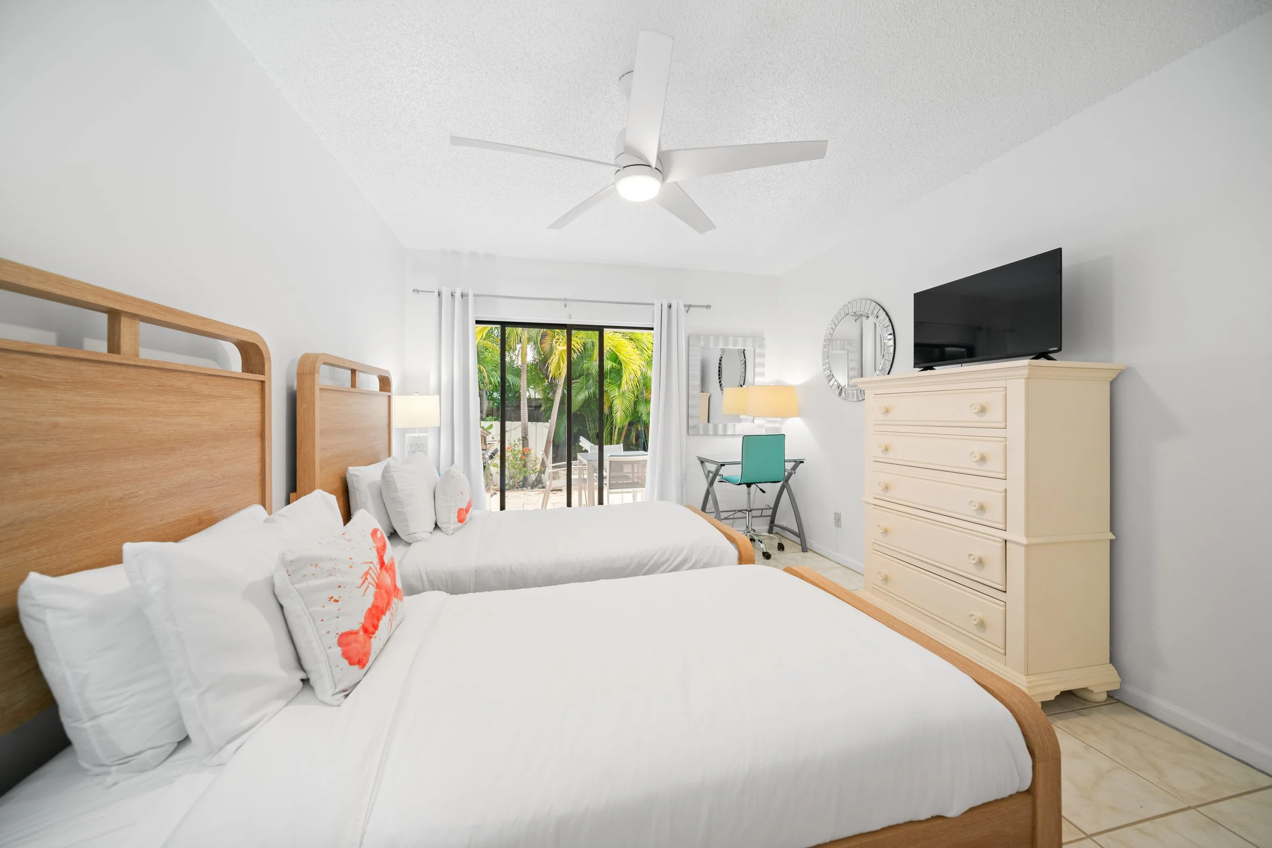 A bedroom with two wooden beds, white bedding, throw pillows with lobster designs, a dresser with a TV on top, a desk with a chair, and a sliding glass door leading to a patio with greenery outside.
