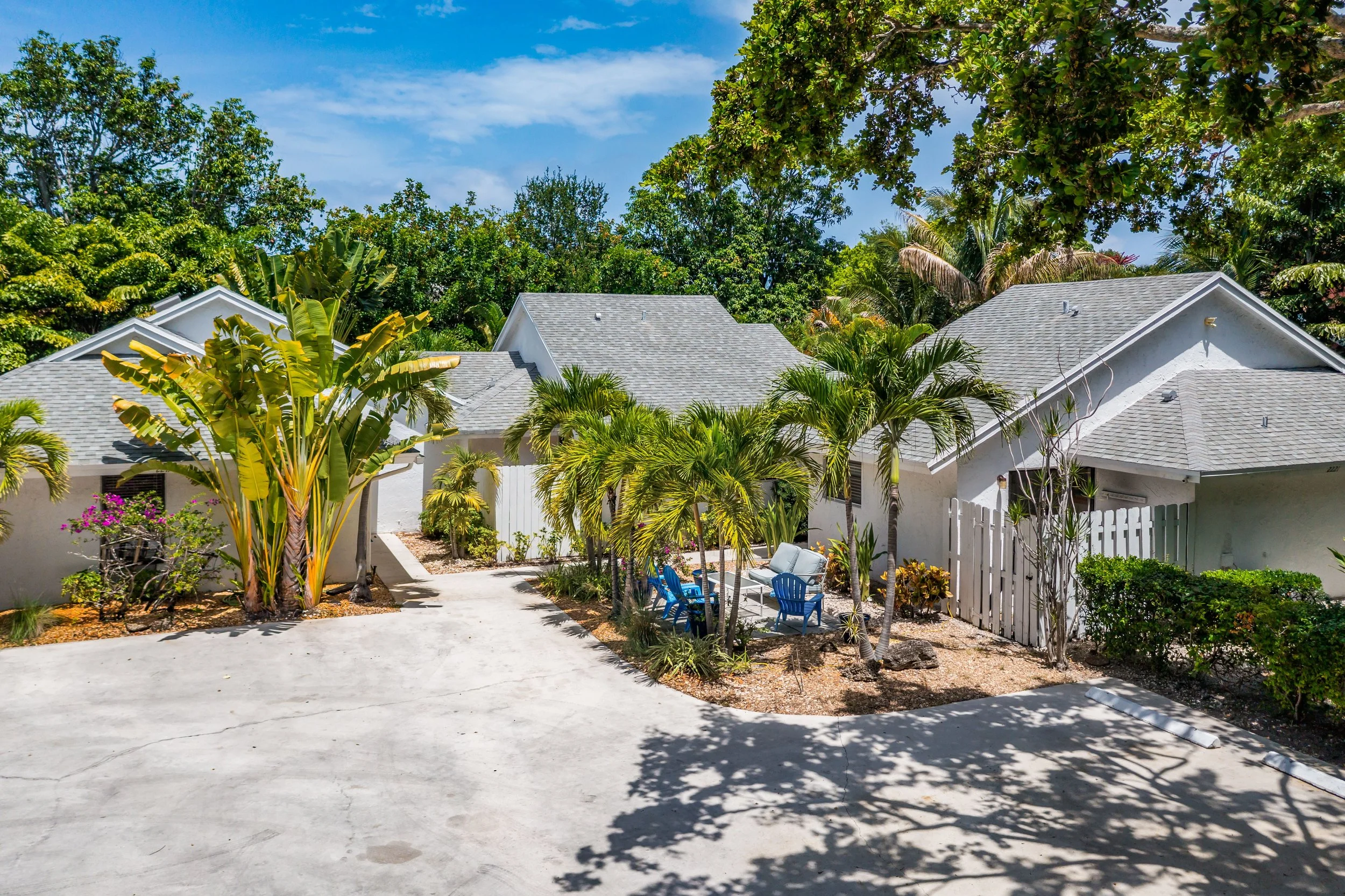 A backyard scene with white houses, gray shingle roofs, and lush green tropical trees and plants. There are blue chairs and a small table on a concrete patio, and a white fence on the right side.
