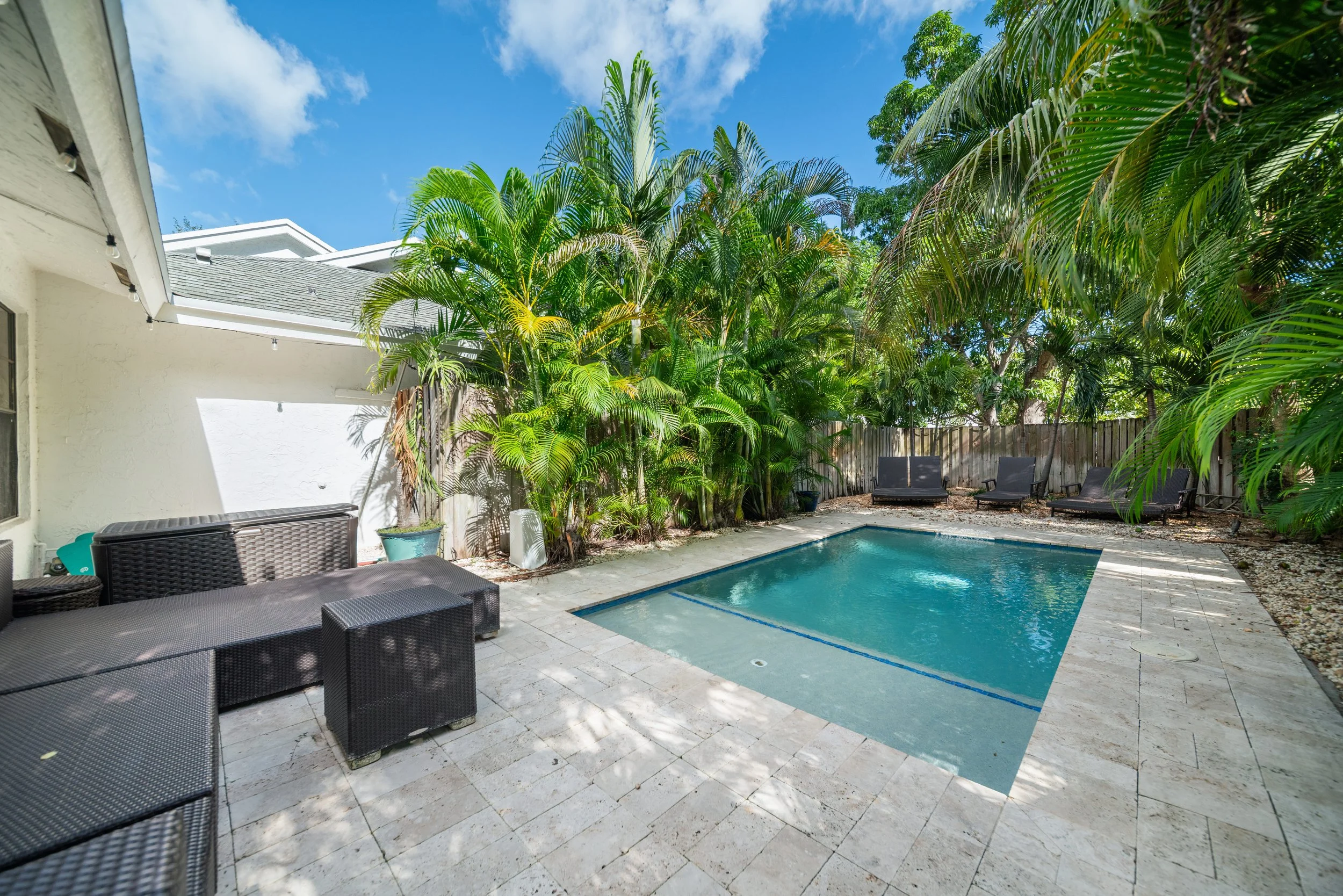 Backyard with a small pool, surrounded by black outdoor furniture and large green palm trees, with a wooden fence and a clear blue sky.
