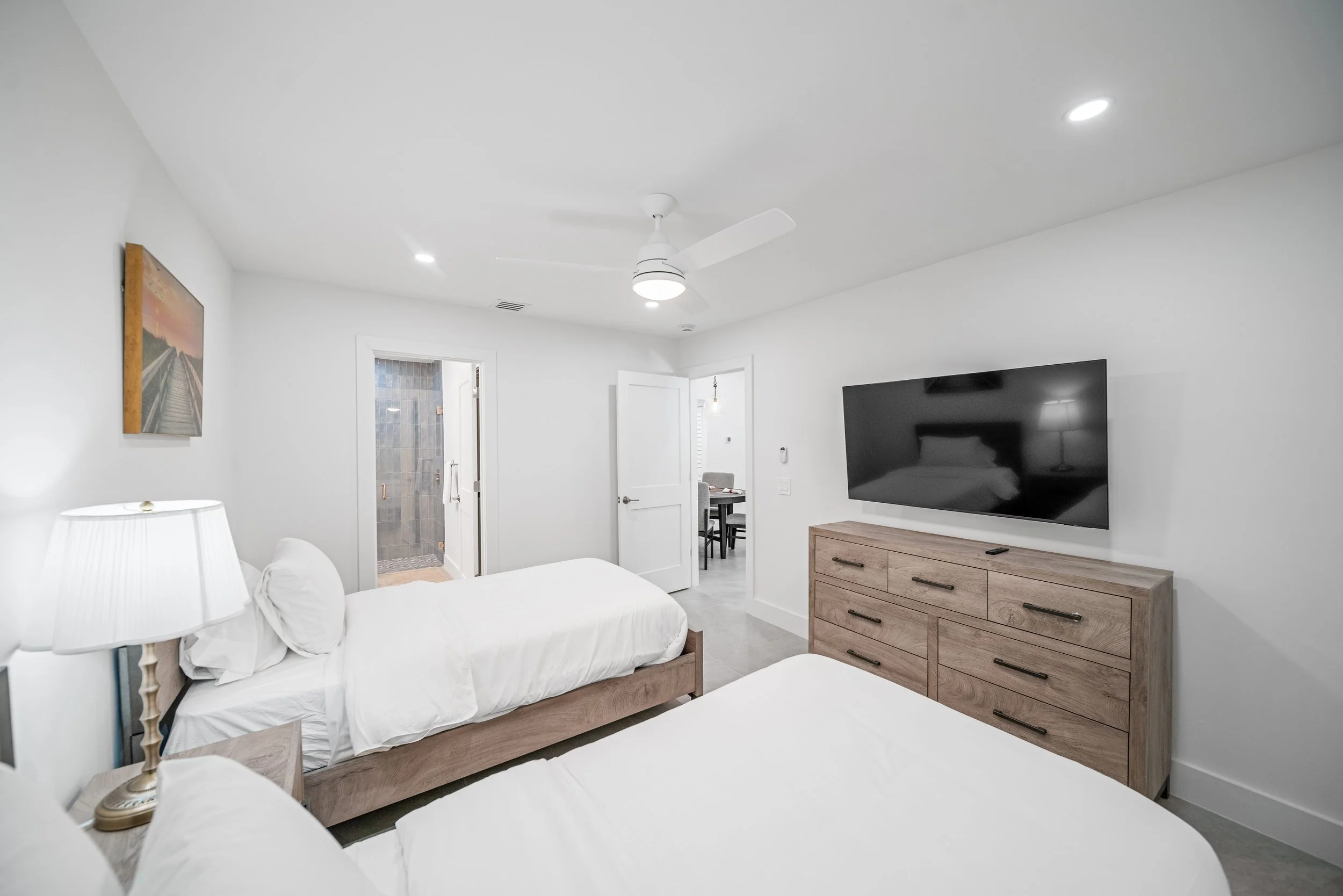 A modern, minimalistic bedroom with white walls and ceiling, two beds with white bedding, a wooden dresser with drawers, a wall-mounted flat-screen TV, a ceiling fan with lights, and a small painting on the wall. There is an open door leading to a ba