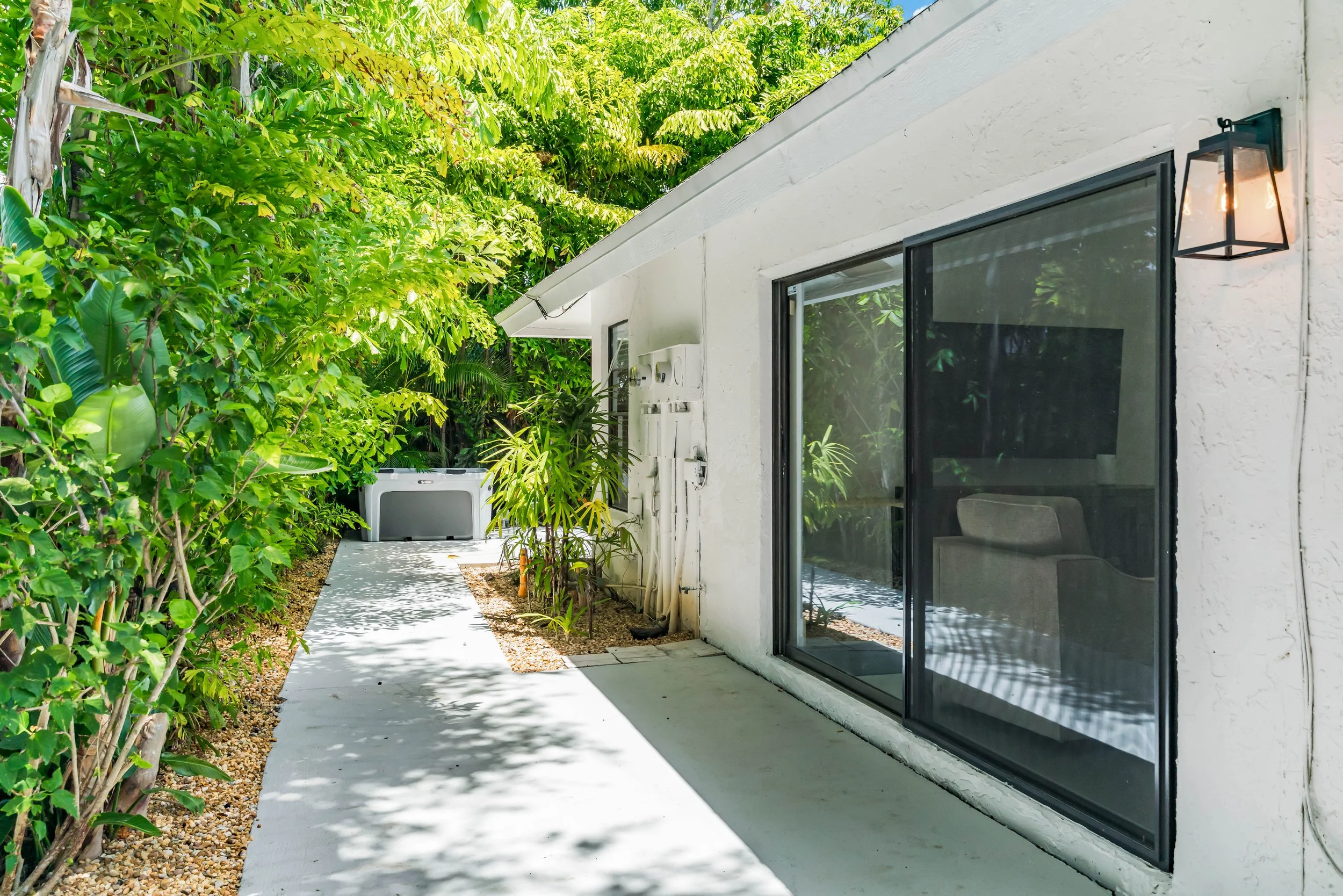 Side view of a white house with large glass sliding door, outdoor lamp, lush green plants, and a concrete patio with gravel border.