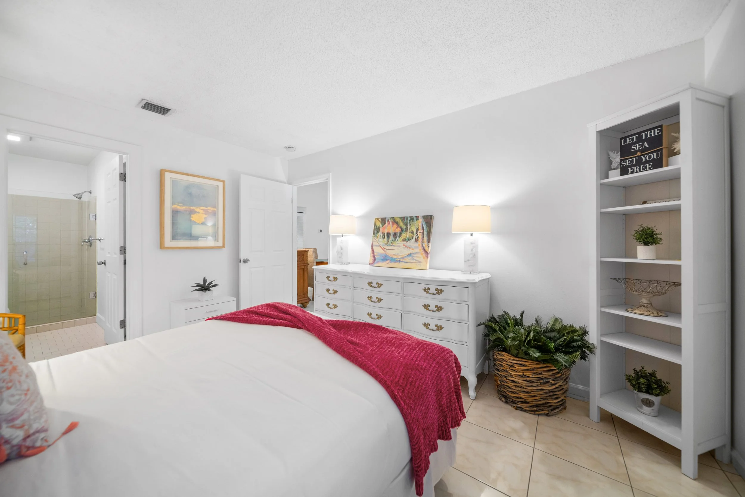 Interior of a bedroom with a white dresser, a colorful painting, two white lamps, and a white bookshelf filled with decorative items and plants. A bed with a red blanket is partially visible in the foreground.