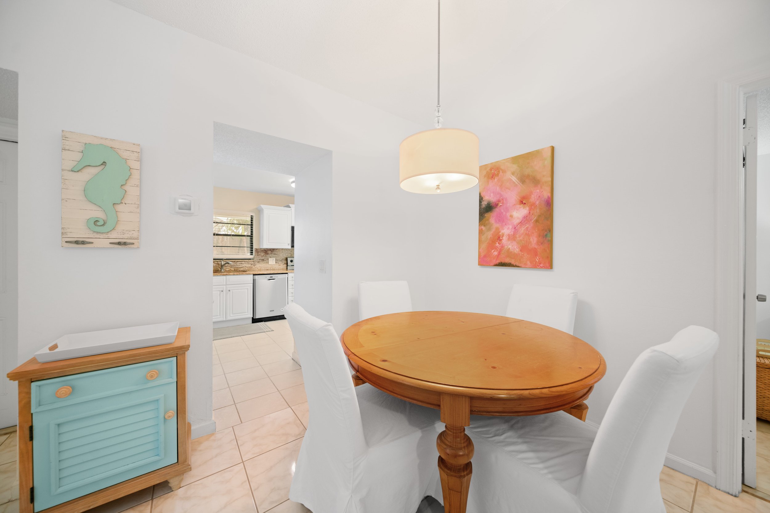 A dining area with a round wooden table and white chairs. Decor includes a seahorse wall art and a large abstract pink and orange painting. The space opens to a kitchen with white cabinets and appliances.