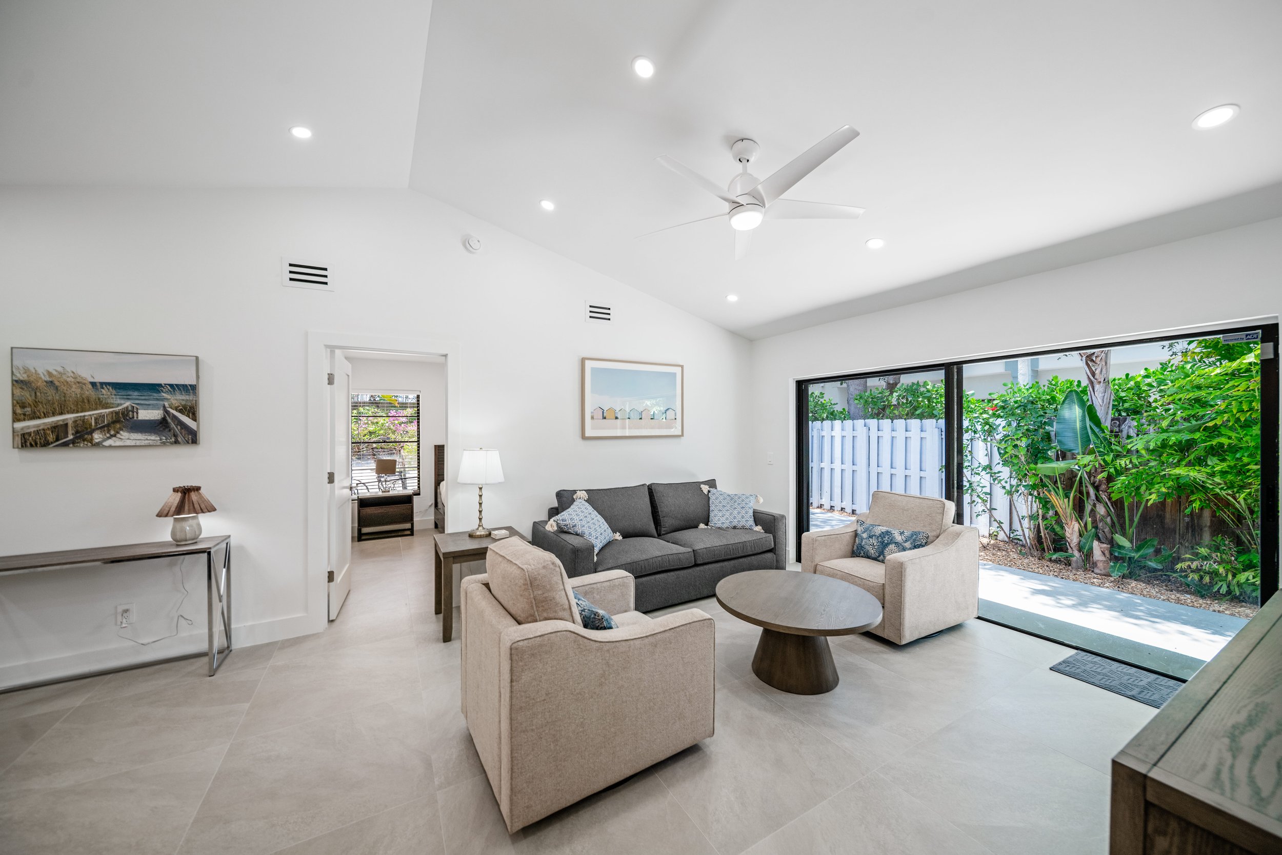 Spacious living room with white walls and ceiling, ceiling fan, large windows with view of green foliage, and comfortable seating including gray and beige sofas and a round wooden coffee table.