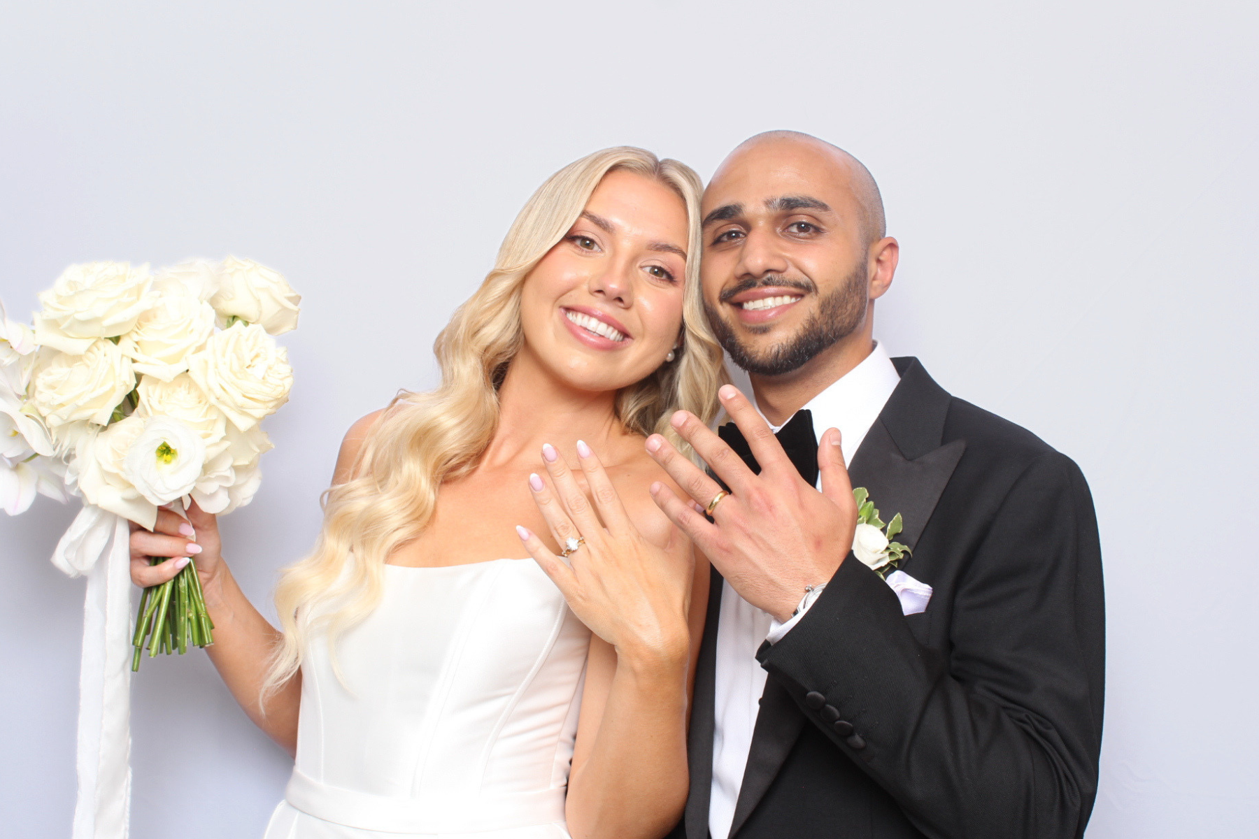 A newlywed couple smiling and showing off their wedding rings. The bride has long blonde hair, a white dress, and holds a bouquet of white roses. The groom has a beard, a shaved head, and wears a black tuxedo with a white shirt and black bow tie. They are standing against a plain white background, close together, displaying their rings.