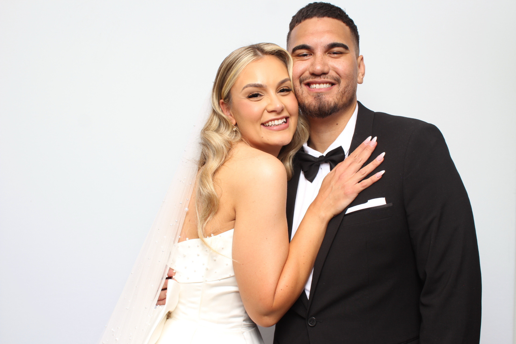 A smiling bride and groom embrace, with the bride in a white wedding dress and the groom in a black tuxedo, against a plain white background.